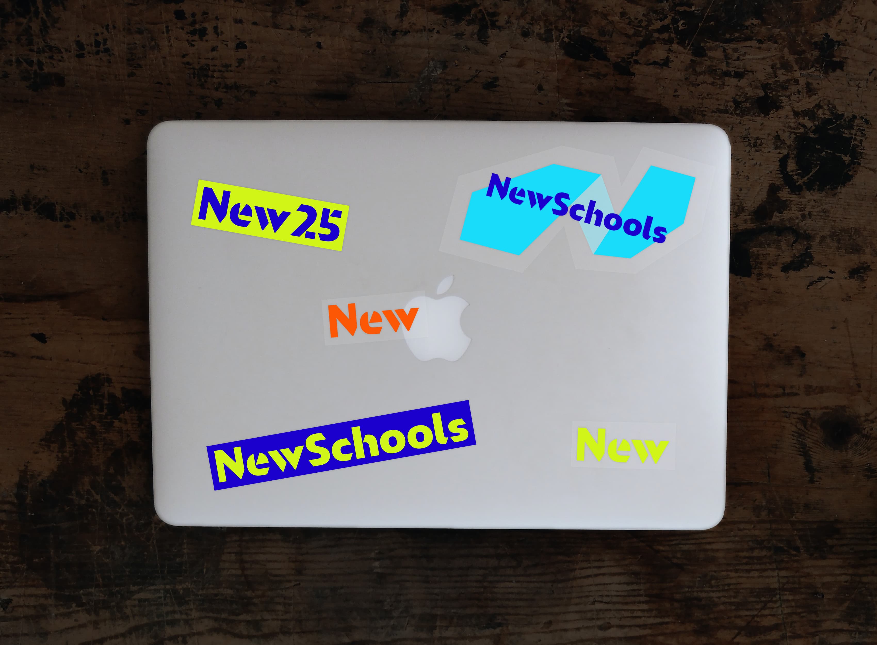 A closed laptop with an Apple logo on its lid sits on a wooden surface. The laptop is covered in several colorful stickers with the words "New25," "NewSchools," and "New" written in various fonts and colors.