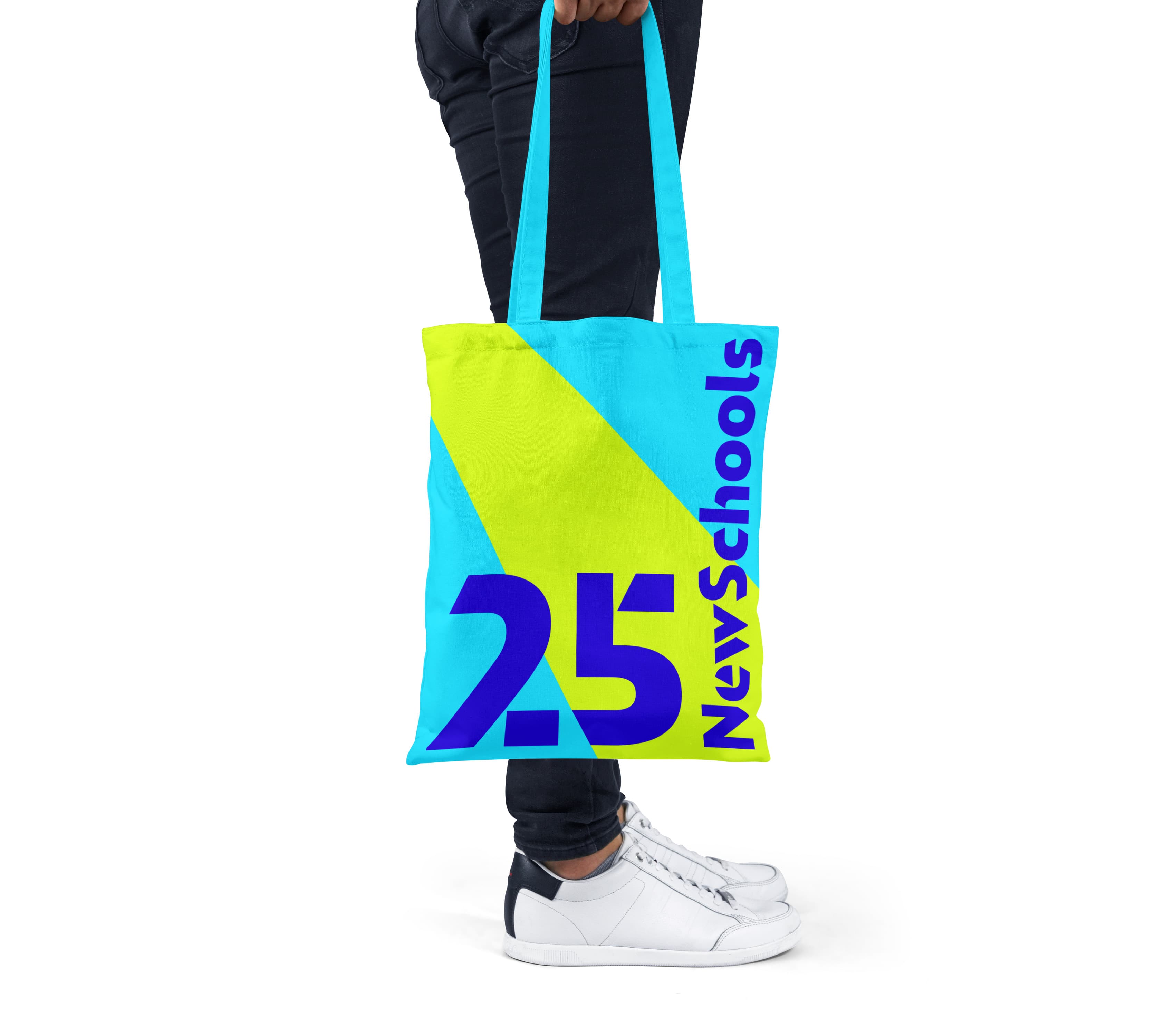 Person holding a bright blue and yellow tote bag with "25 NewSchools" written on it in bold purple and blue text, standing against a plain background. The person wears dark pants and white sneakers.