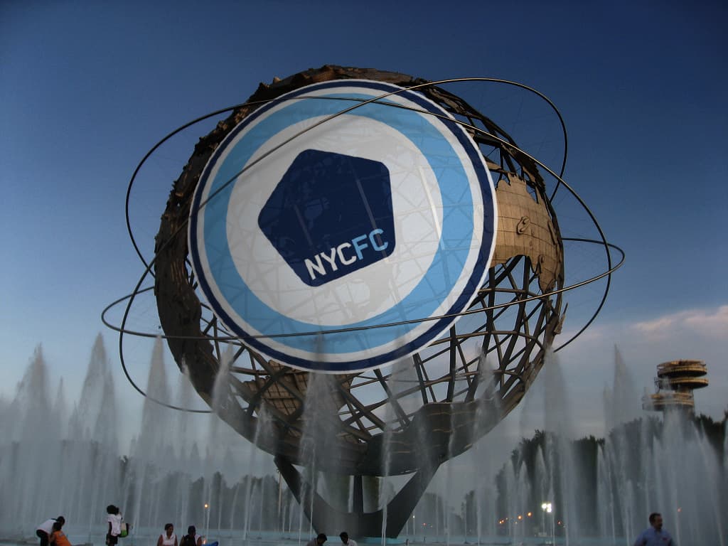 A large spherical metal structure resembling a globe, situated in the middle of fountains at dusk. The globe is overlaid with a blue and white NY City FC (NYCFC) logo in the center. People are seen walking and sitting around the fountain area.