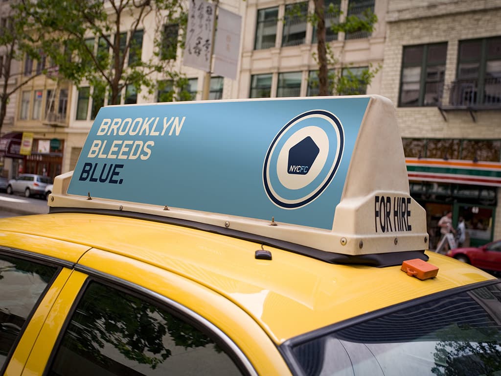 A yellow taxi cab in an urban setting features an advertisement on its roof that reads, "BROOKLYN BLEEDS BLUE," with branding for WHC. The sign is blue and white, situated in front of a beige building with trees and other storefronts visible in the background.