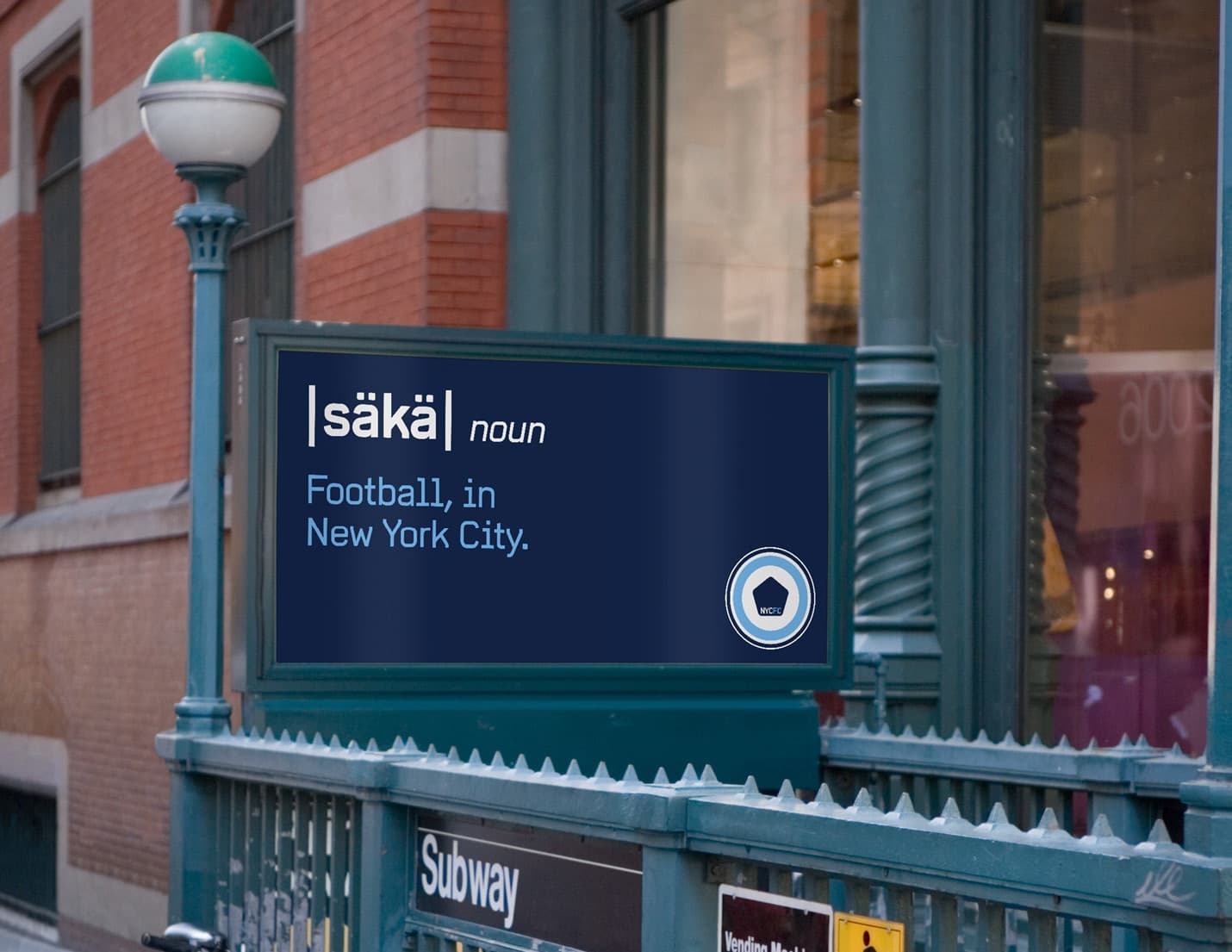 A digital sign near a subway entrance in an urban setting displays the text: "säkə noun Football, in New York City." The sign has a dark blue background with white letters, and the subway entrance is surrounded by buildings with brick walls and large windows.
