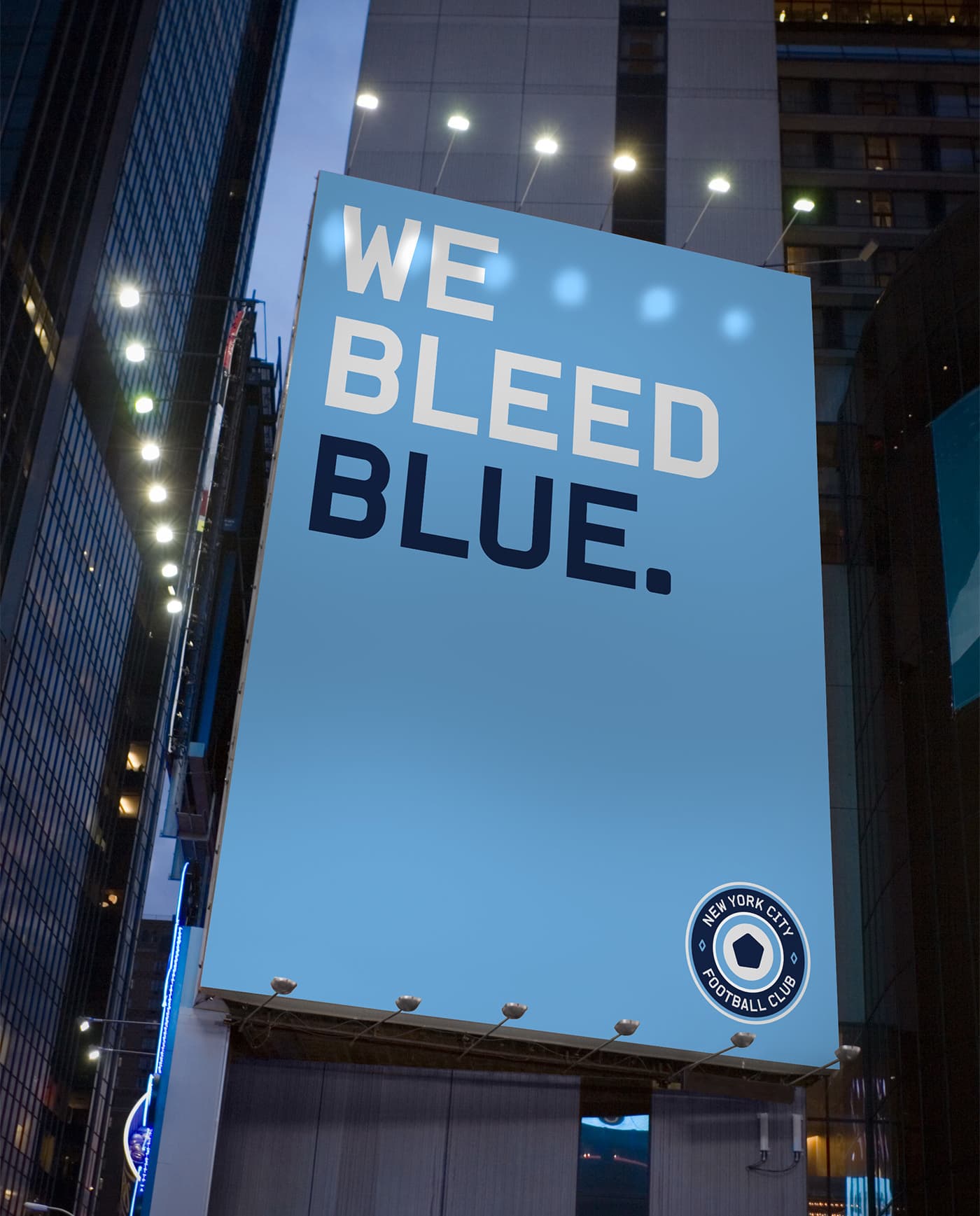 A large, illuminated billboard on a city street at night displays the message "WE BLEED BLUE." in bold white and blue text against a light blue background. The New York City Football Club logo is at the bottom right corner of the billboard.