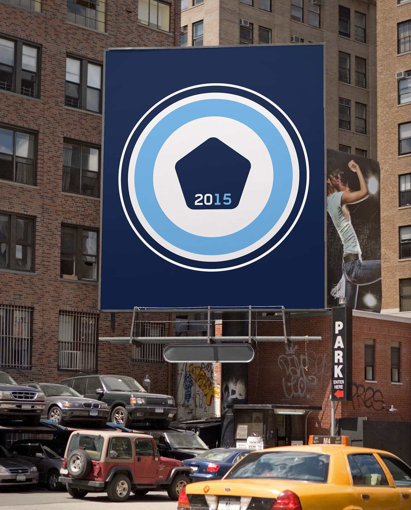A large billboard with a blue and white emblem featuring the year "2015" is displayed on the side of a brick building in an urban area. Cars are parked below the billboard, and buildings with various wall art and graffiti are visible in the background.
