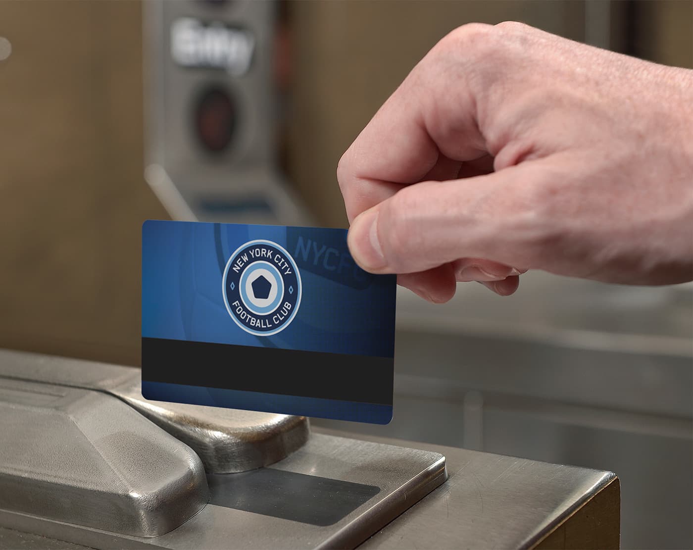 A close-up of a hand holding a New York City Football Club card, scanning it at a turnstile. The card features the club's emblem and a blue background with a black stripe. The scanning device and a blurred background are partially visible.