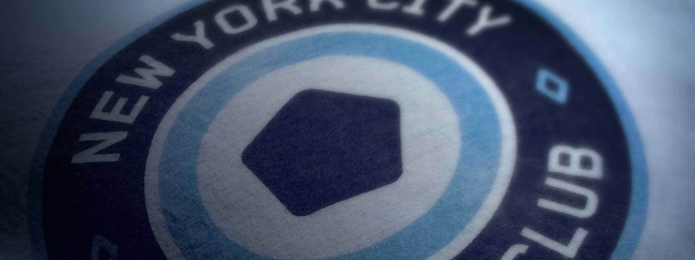 Close-up of the New York City Football Club logo featuring a stylized circular emblem with "New York City" written on the top half and "Club" on the bottom. The interior hosts a blue hexagon surrounded by light blue and dark blue rings.
