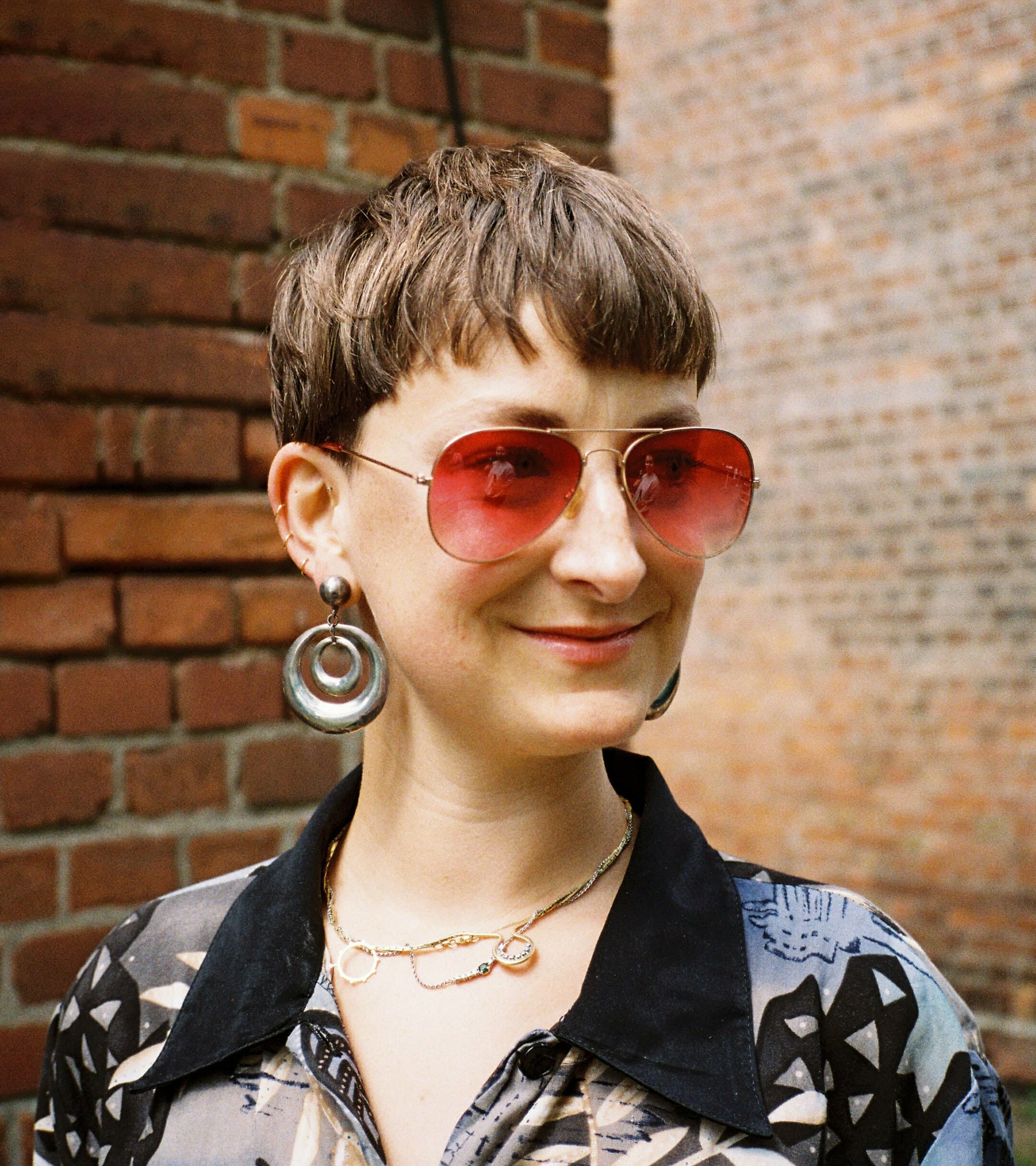 A person with short hair and wearing large circular earrings stands outside in front of a brick wall. They are wearing red-tinted aviator sunglasses and a patterned shirt, and they are smiling slightly.