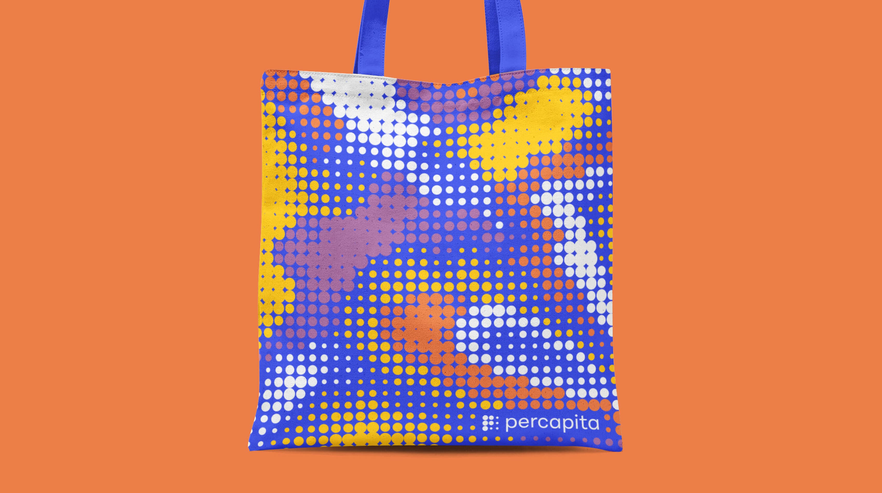 A tote bag with a colorful pixelated design in yellow, blue, purple, and white dots on an orange background. The bag has blue handles and the brand name "percapita" is printed in white at the bottom right corner.