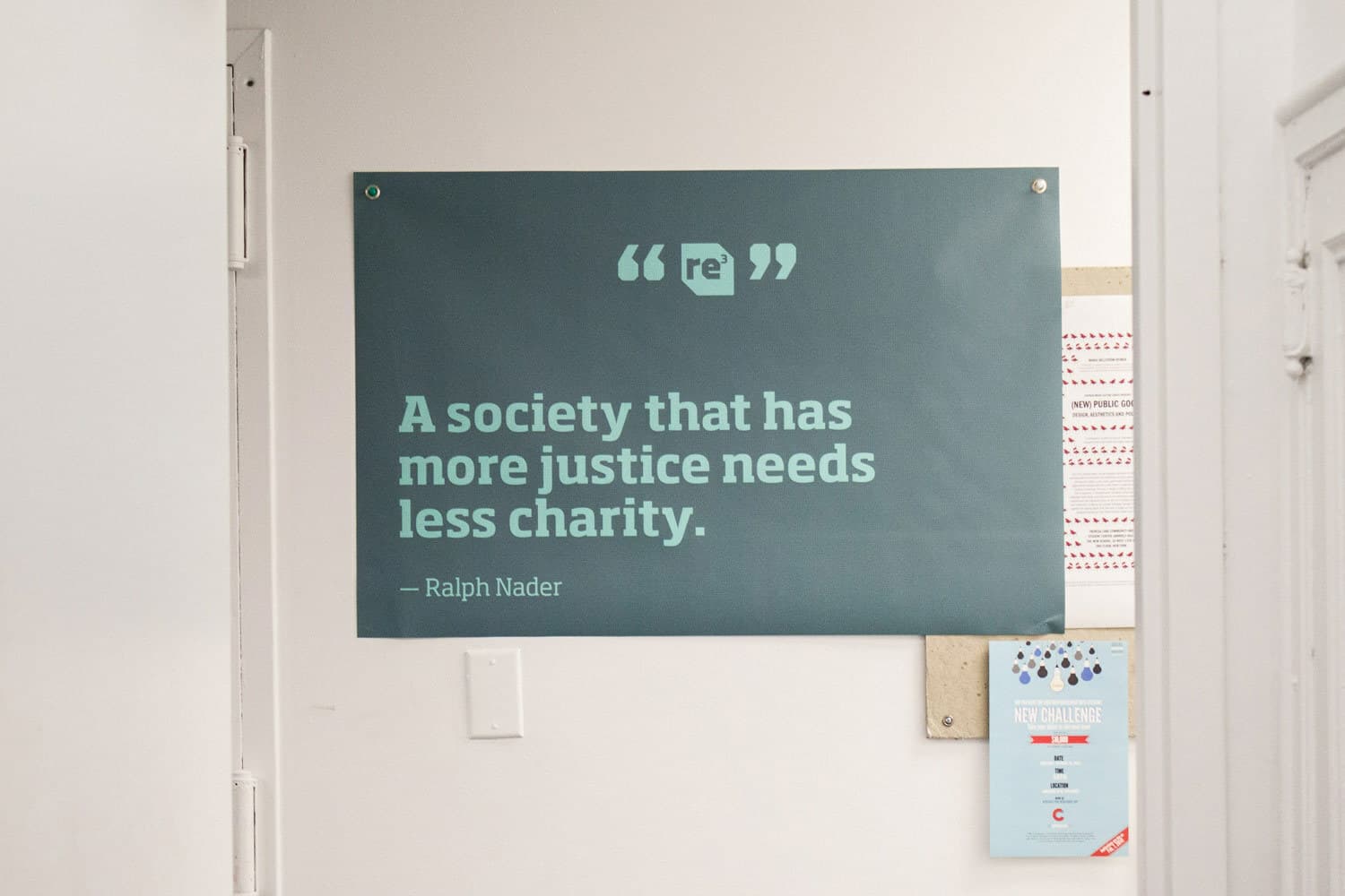 A poster pinned to a white wall reads, "A society that has more justice needs less charity. - Ralph Nader." Below the quote, there is a small logo with "re" in the center. A calendar and other papers are partially visible on the wall to the right.