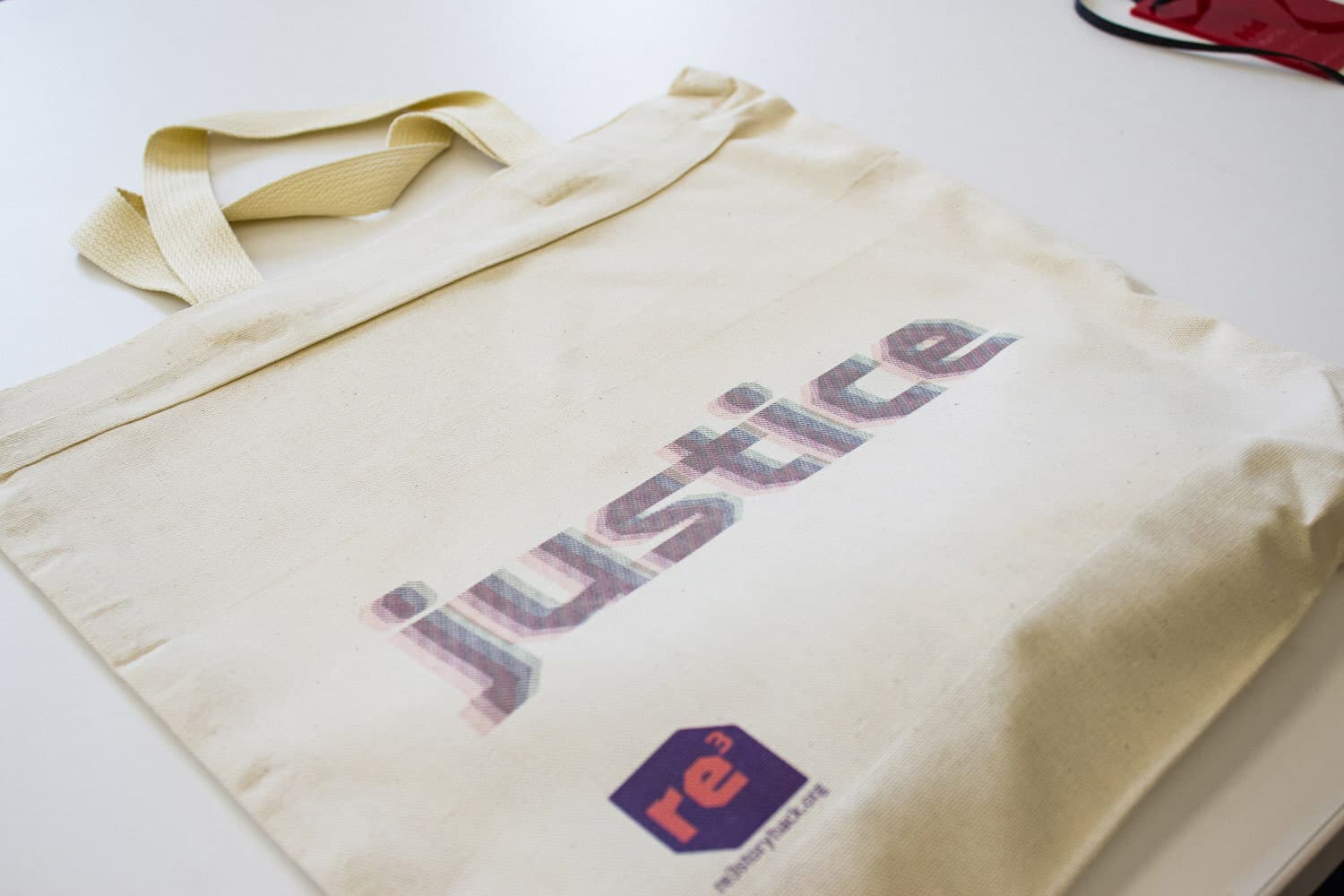 A beige tote bag lies on a white surface. The word "justice" is printed in large, colorful letters on the front of the bag. Below it, there is a small purple and red logo with the text "re3," followed by smaller text that is not clearly legible.