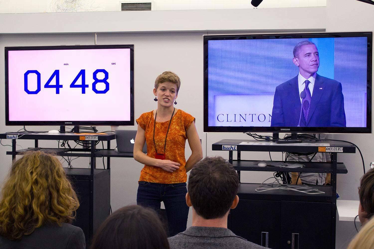 A woman in an orange shirt stands and speaks in front of an audience. Behind her, two screens are visible; the left screen shows a timer at "04:48," and the right screen displays a man in a suit speaking. The room appears to be a conference or meeting setting.