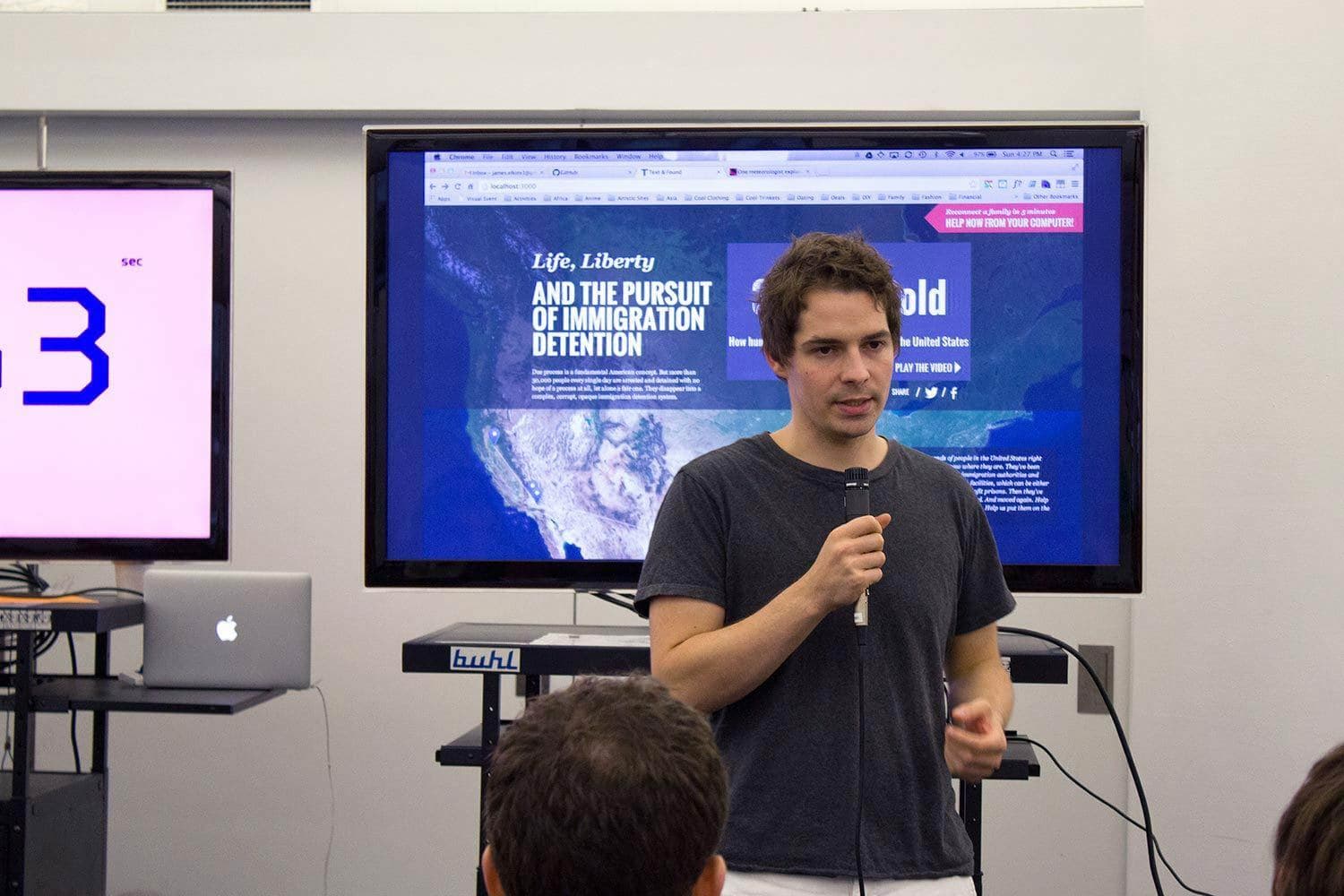 A man in a dark T-shirt speaks into a microphone in a room, with people seated before him. Behind him, there is a screen displaying an article titled "Life, Liberty, AND THE PURSUIT OF IMMIGRATION DETENTION." A laptop is visible on a table to his right.