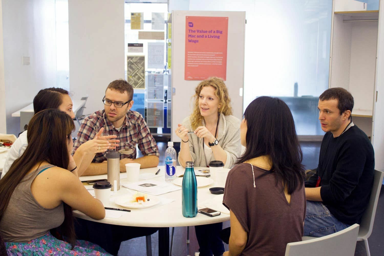 A group of six people engaged in a discussion around a table in a bright, modern room. Various items, including cups, a notebook, and a bottle, are on the table. A poster titled "The Value of a Big Mac and a Living Wage" is visible in the background.