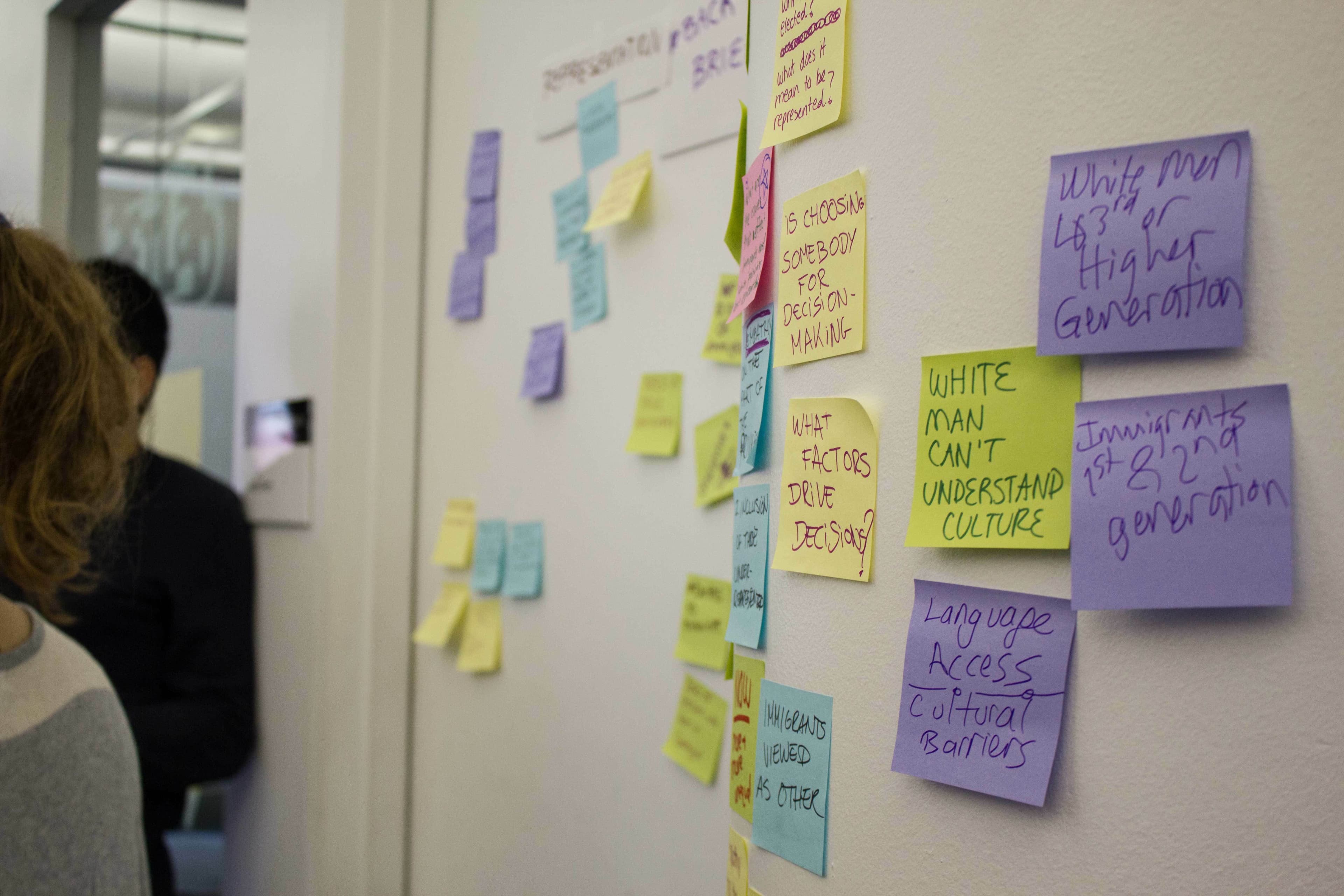 A wall covered with various colorful sticky notes, each containing written phrases and ideas related to understanding culture. Some notes read "White man can't understand culture" and "Immigrants 1st & 2nd generation." A person is partially visible in the foreground.