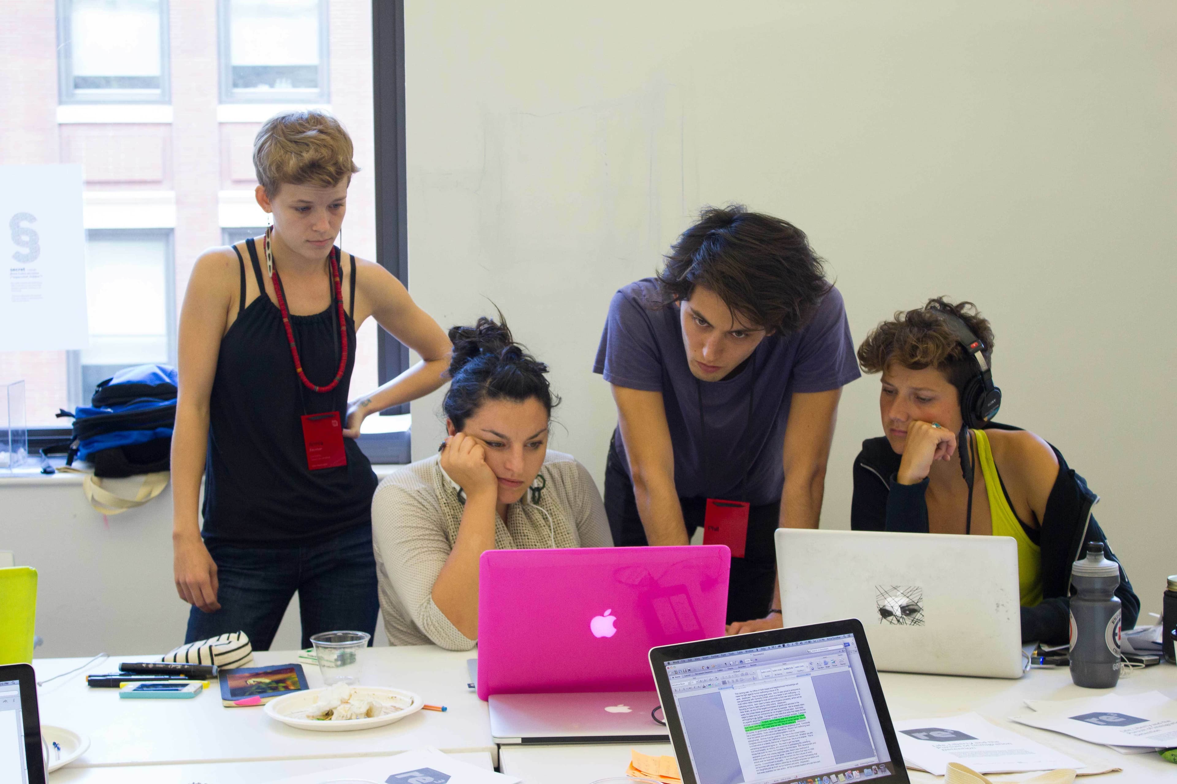 Four individuals are gathered around a table, each using a laptop. The person with a pink laptop appears focused, while the others look on attentively. Various papers, bowls, and office supplies are scattered on the table. A window in the background lets in natural light.
