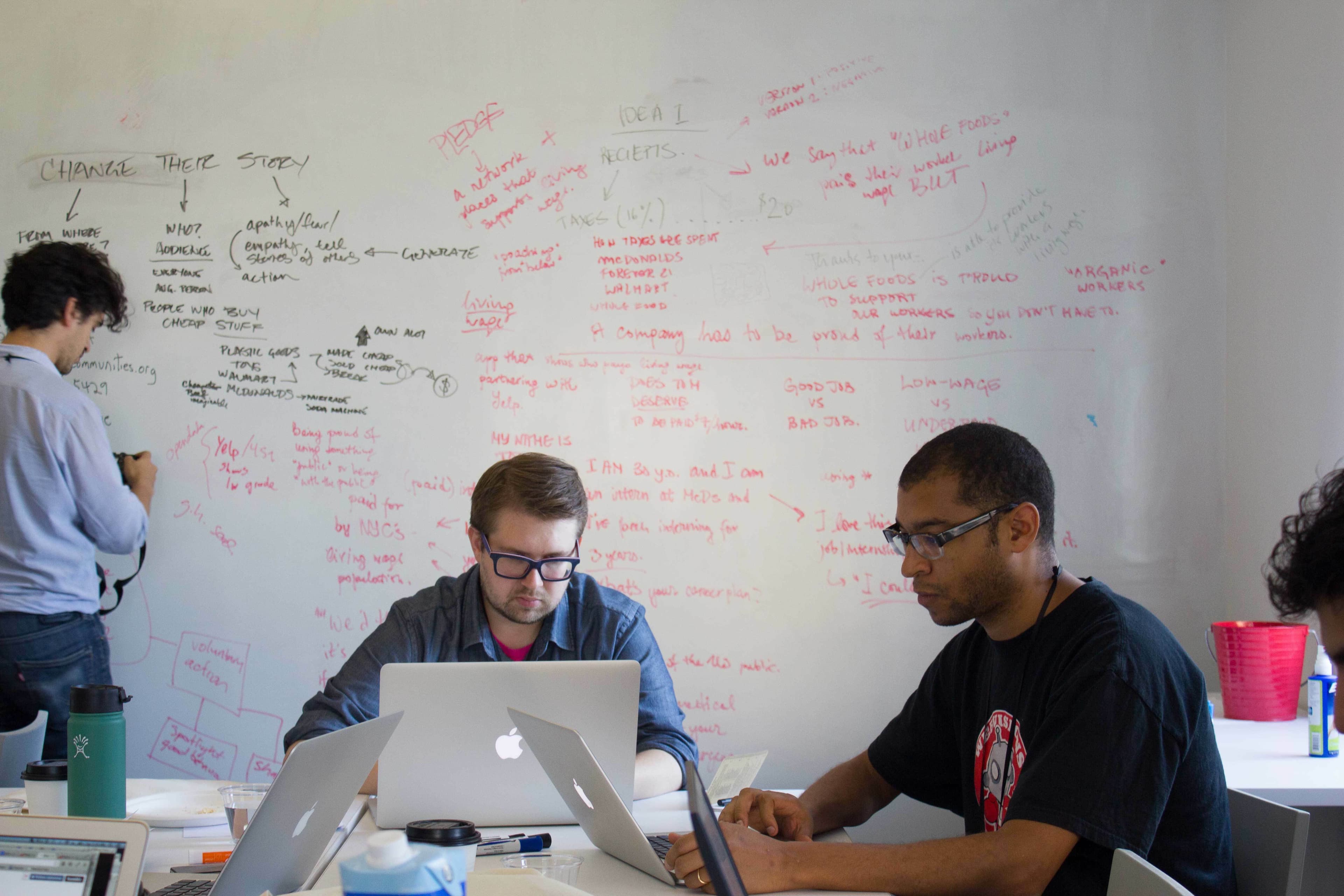 A group of people work in a room with whiteboards covered in handwritten notes, diagrams, and ideas. Two people are seated at a table engrossed in their laptops, while another is standing and writing on the whiteboard. Various items like coffee cups are on the table.