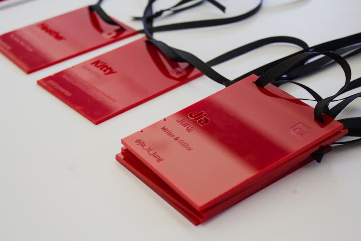 Close-up of three red name badges on a table. The badges have black lanyards attached. The nearest badge reads, "Jia, Writer & Editor, @Jia_J_Jung" in white text. The other two badges behind it are partially visible, showing different names and titles.