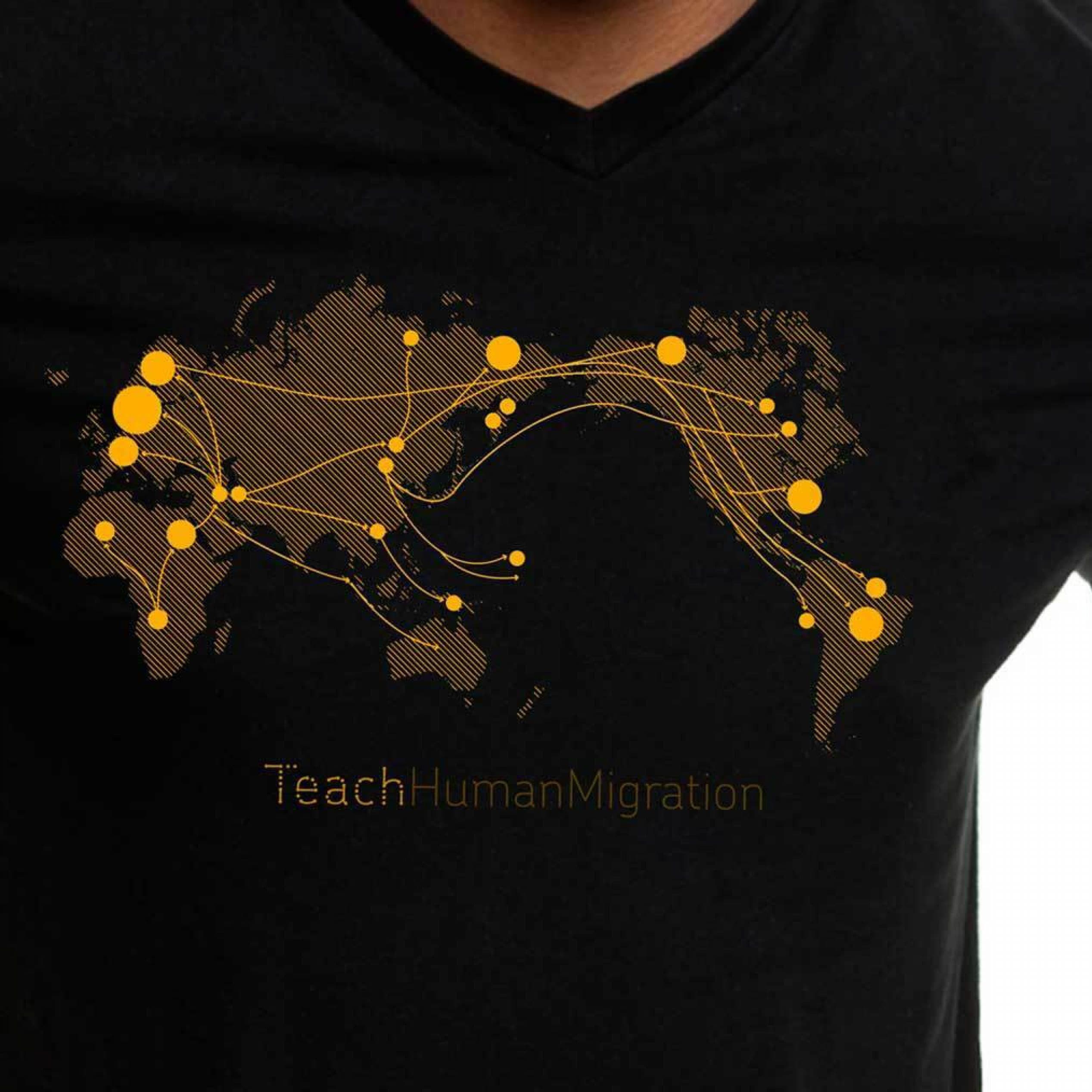 A person wearing a black V-neck T-shirt featuring a stylized world map. The map depicts migration routes with yellow lines and dots connecting various global locations. The text "TeachHumanMigration" is printed below the map in yellow.