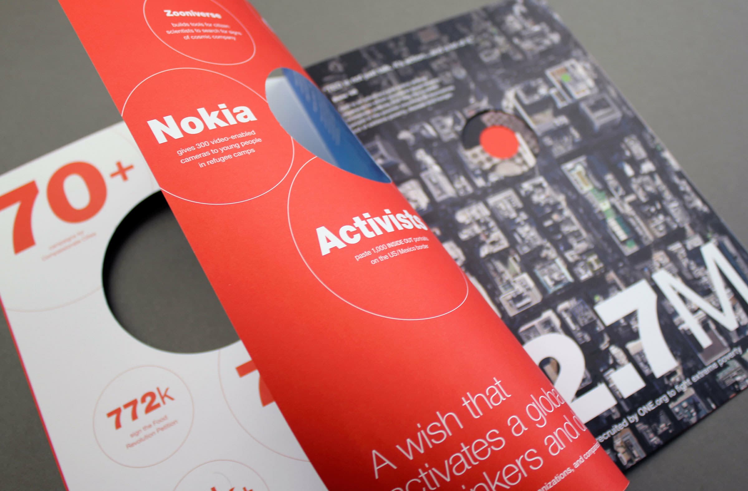 A brochure with a grey and red color scheme is open. Visible text includes "Activistas," "Nokia," "70+," "772k," and "2.7M." One page has a circular cutout revealing part of a diagram or map beneath.