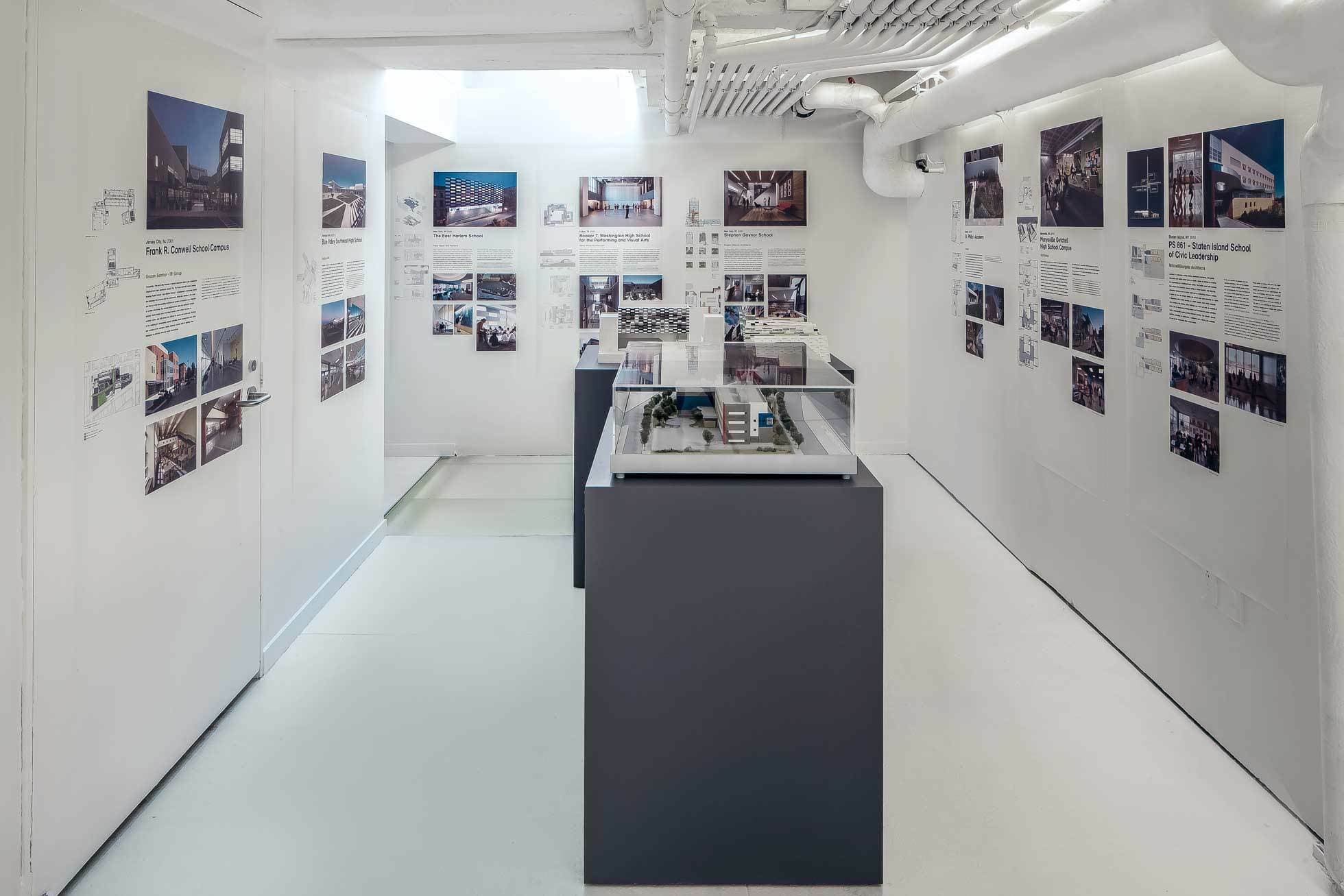 A modern exhibition space features white walls adorned with architectural posters and images. Several glass-topped display cases containing detailed building models and blueprints are positioned along the center of the room. The ceiling has exposed pipes.