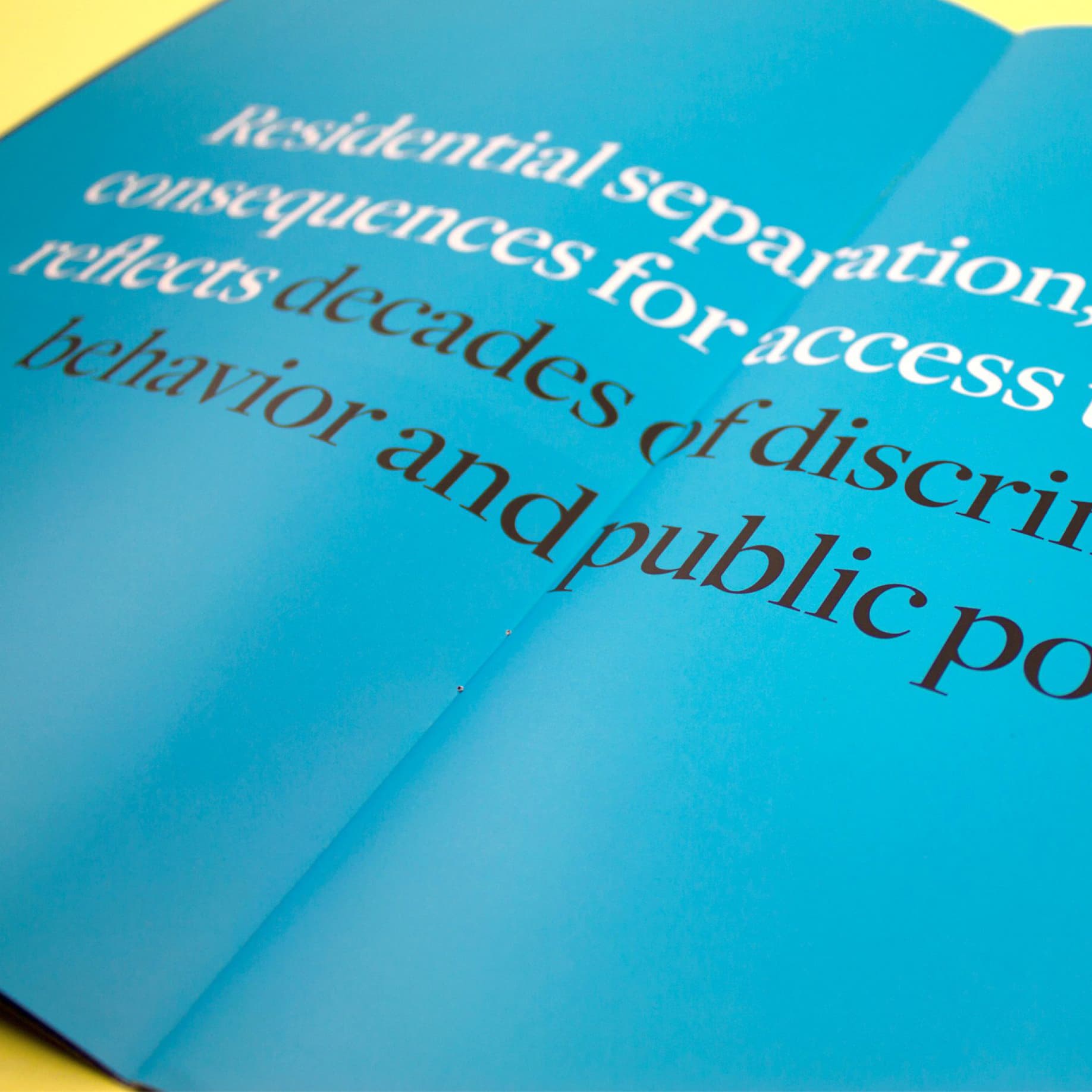 A close-up of an open book or magazine with a blue page and white text. Some black text is also visible. The text discusses residential separation, consequences for access, decades of discrimination, behavior, and public policy. The text spans the book's crease.