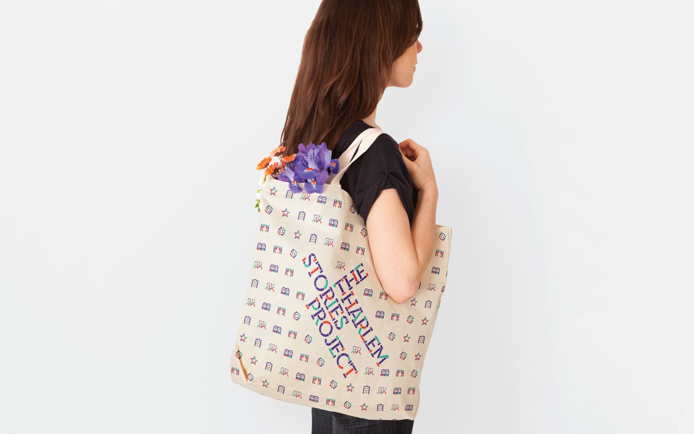 A woman with long brown hair is standing sideways, holding a beige tote bag over her shoulder. The bag has colorful text that reads "THE TILE HEALING PROJECT" and is filled with purple and orange flowers. The background is plain and white.