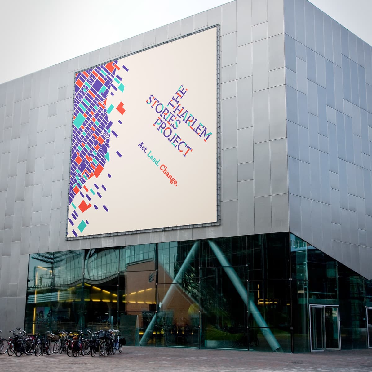 A large building facade displays a colorful billboard for the "Harlem Stories Project" with the tagline "Act. Lead. Change." The billboard features a geometric, multicolored map. Bicycles are parked in front of the building beside large glass windows.