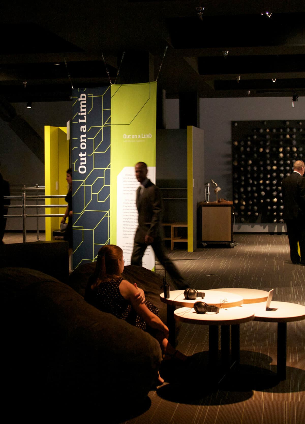 A dimly lit interior with people interacting in a modern exhibit space. A green sign reads "Out on a Limb." Two white tables with headphones and other objects are in the foreground. A large black wall installation is visible in the background.