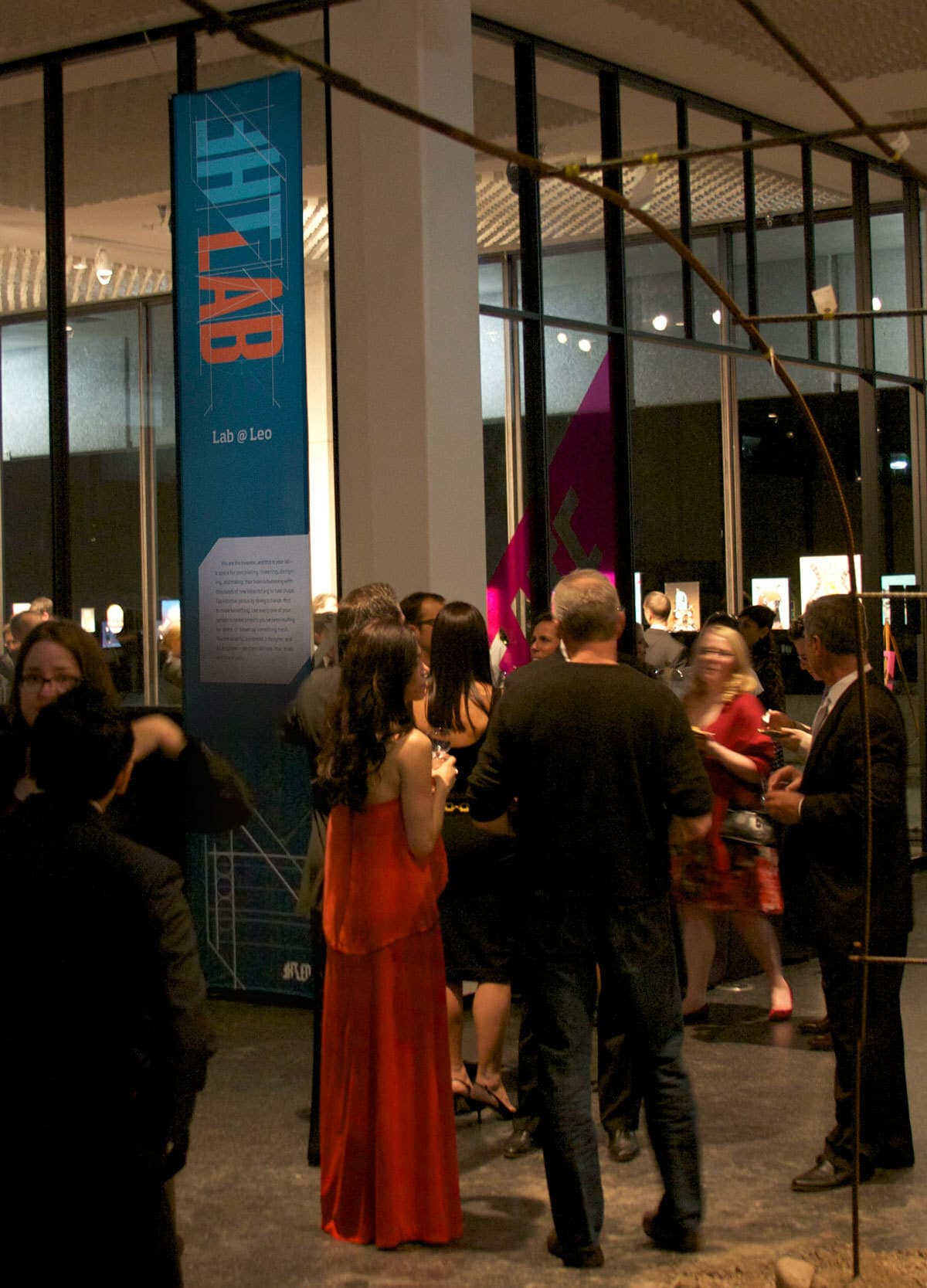 A group of people socializing at an indoor event near a tall sign with "LAB" written vertically on it. Guests are dressed in semi-formal attire, including a woman in a red dress and a man in a suit. The background features large windows and exhibit displays.