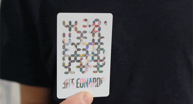 A person holds a white card with colorful, reflective, hexagonal patterns and the text "THELEONARD" at the bottom. The card appears to be turned over, showing a different set of patterns. The background is a plain, dark-colored shirt.