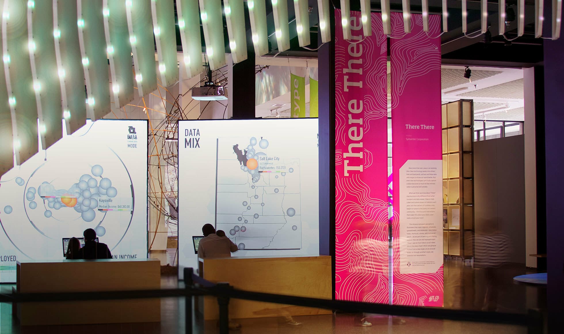 A modern exhibition space featuring bright, interactive screens and panels. Two people are viewing illuminated data visualizations, maps, and large pink informational banners with wavy patterns. The room has a contemporary design with overhead lighting accents.