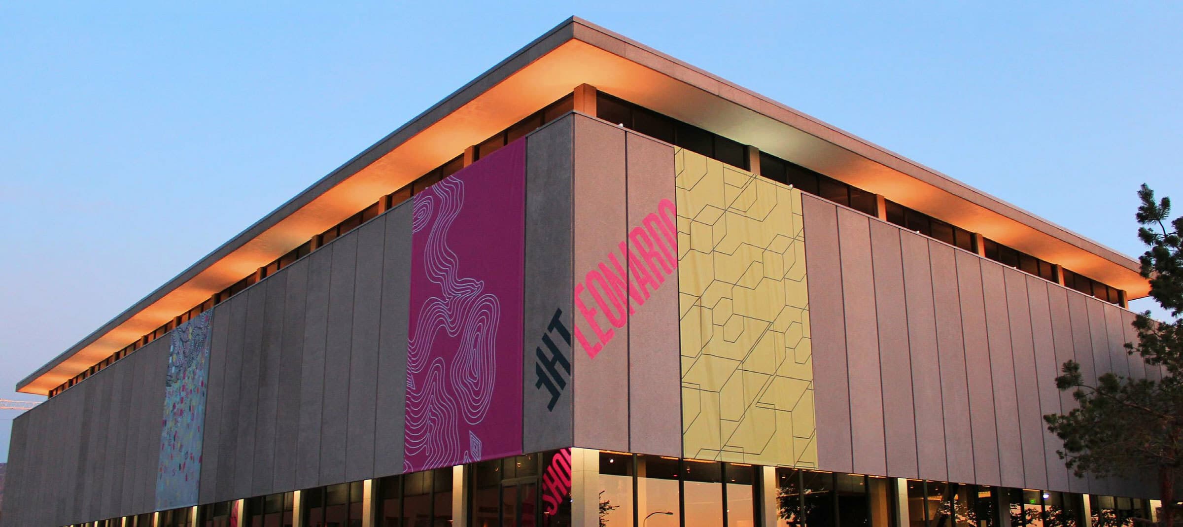 A modern building with a flat roof and cut-out windows near the top, adorned with colorful vertical banners. The left banner is pink with abstract white lines, and the right banner is yellow with angular patterns. The building is lit by warm lights at dusk.