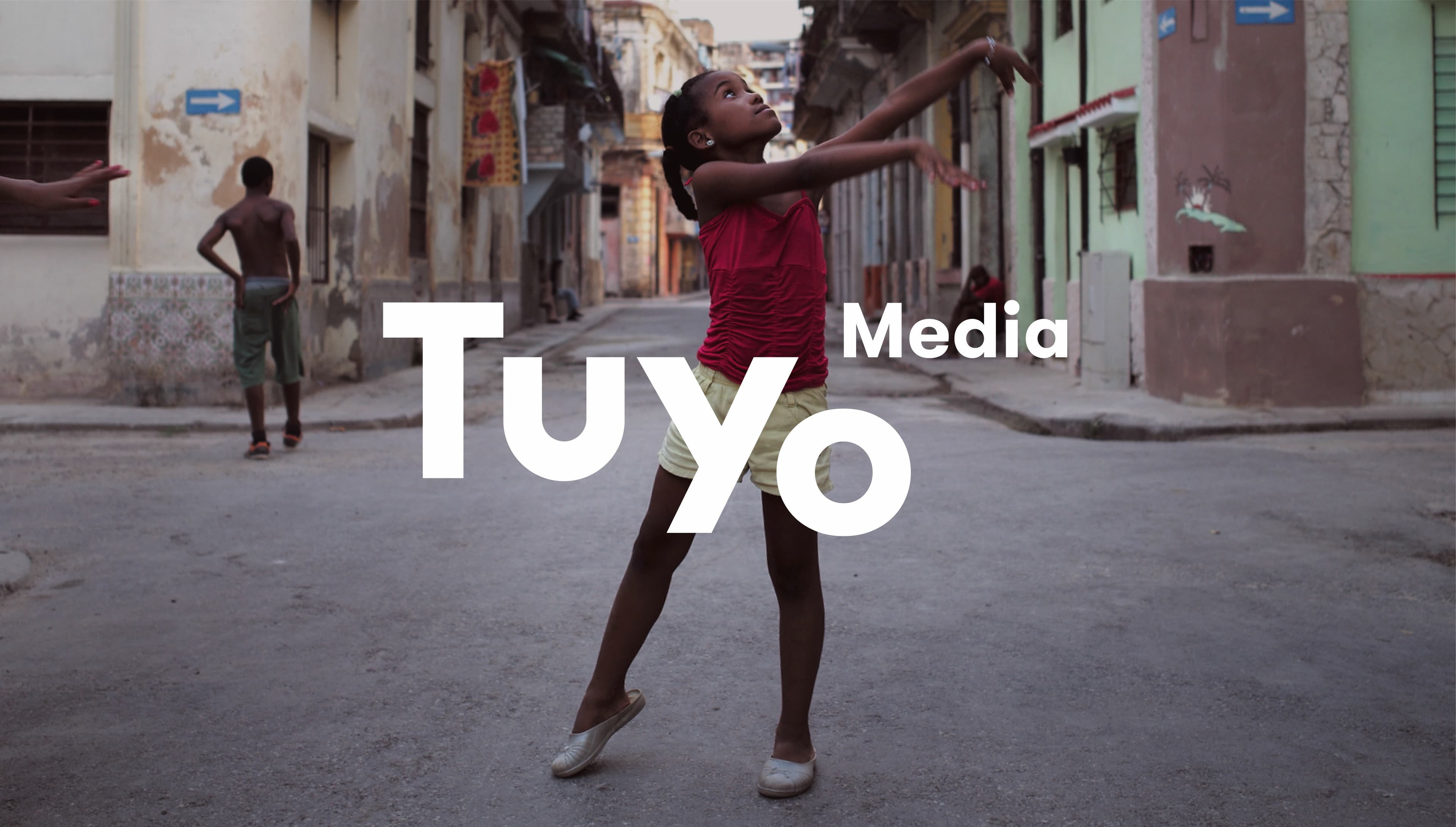 A young girl in a red top and yellow shorts dances barefoot on an empty narrow street lined with colorful, worn buildings. A boy stands in the background with his back turned. The words "Tuyo Media" are overlaid on the image.
