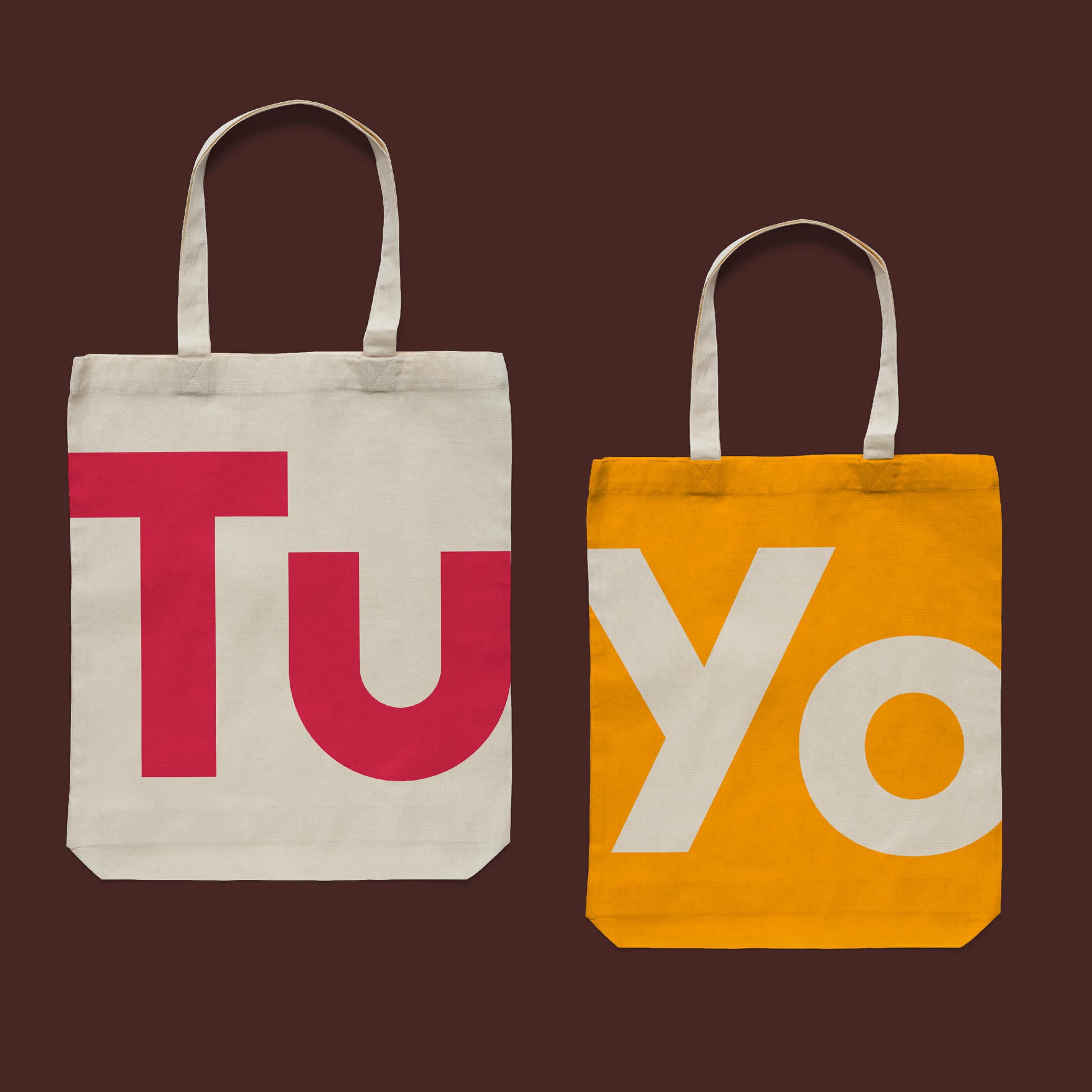 Two tote bags are placed side by side against a dark brown background. The left bag is beige with large red letters "T" and "u". The right bag is orange with large white letters "Y" and "o". Both bags have long handles and a minimalist design.