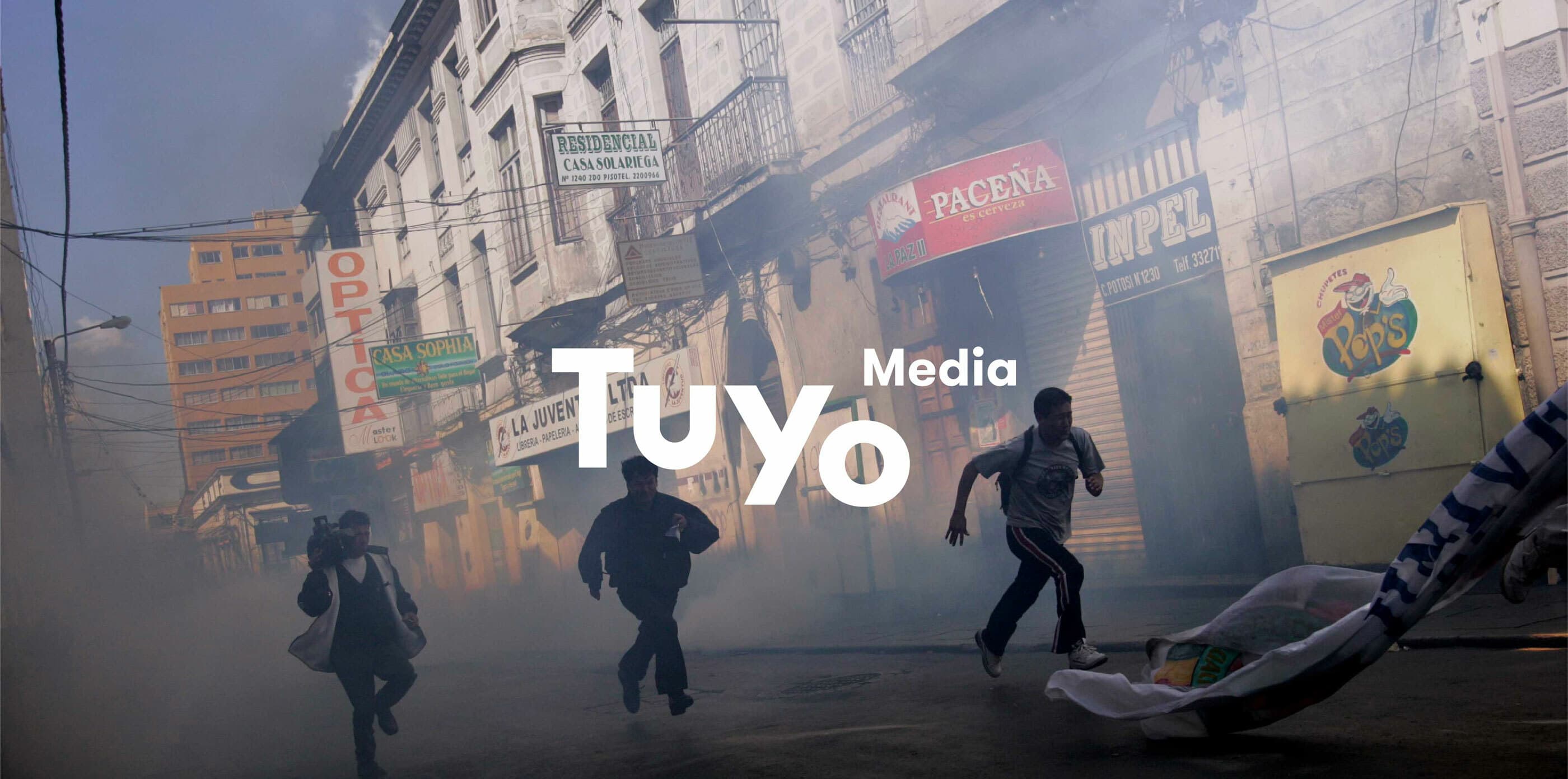 People are running down a smoke-filled street lined with buildings and shop signs. The street appears to be in turmoil. In the foreground, three individuals are actively moving away from the smoke. White text overlay reads "Tuyo Media.