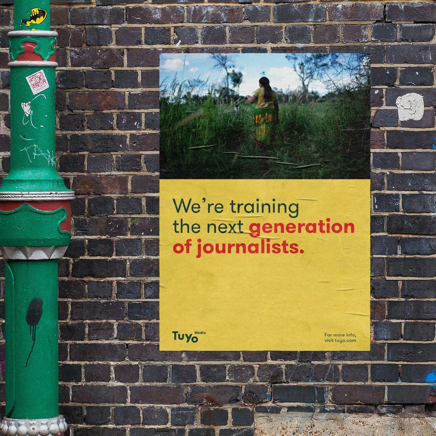 A poster on a brick wall shows an image of a person in a field. The text reads, "We’re training the next generation of journalists." Below is the logo for Tuyo along with a note for more information. A decorated green pole is partially visible on the left.