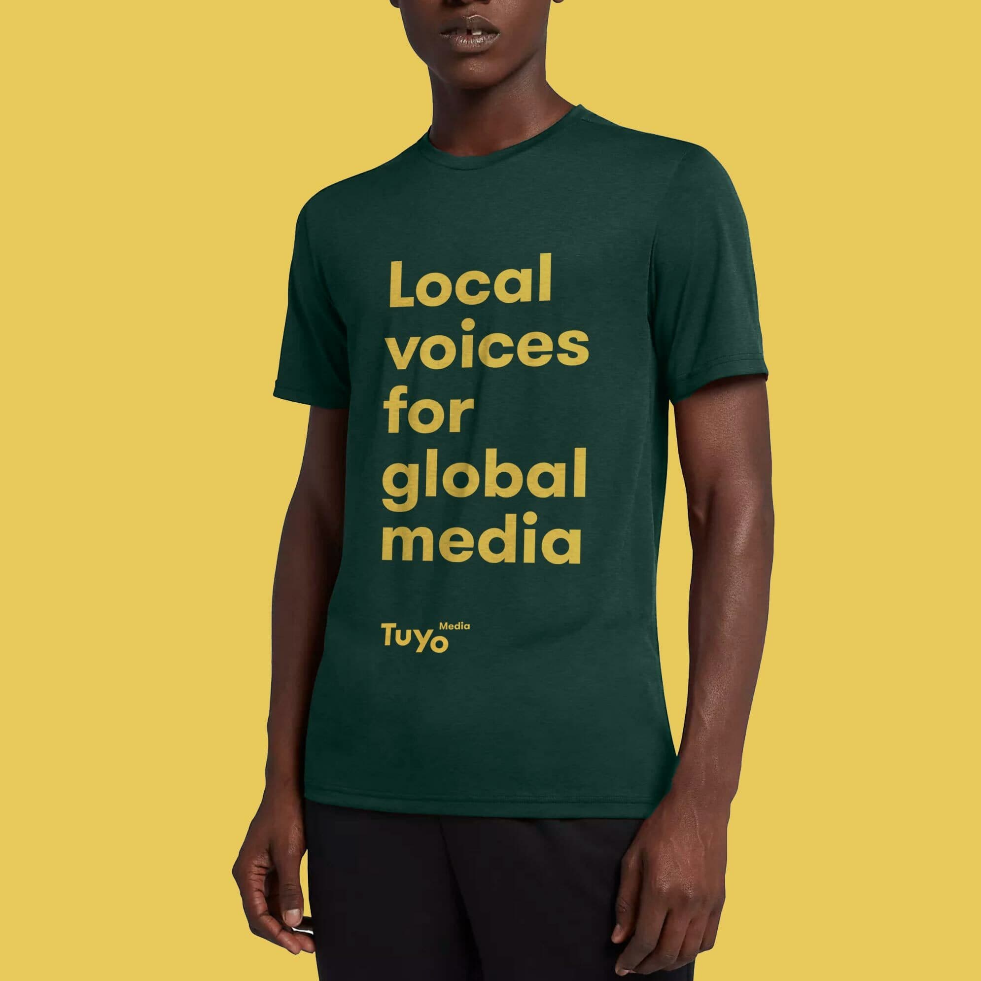 A person wearing a dark green t-shirt with the words "Local voices for global media" and the logo "Tuyo Media" in yellow text. The background is a solid yellow color. The person is standing with one arm by their side.