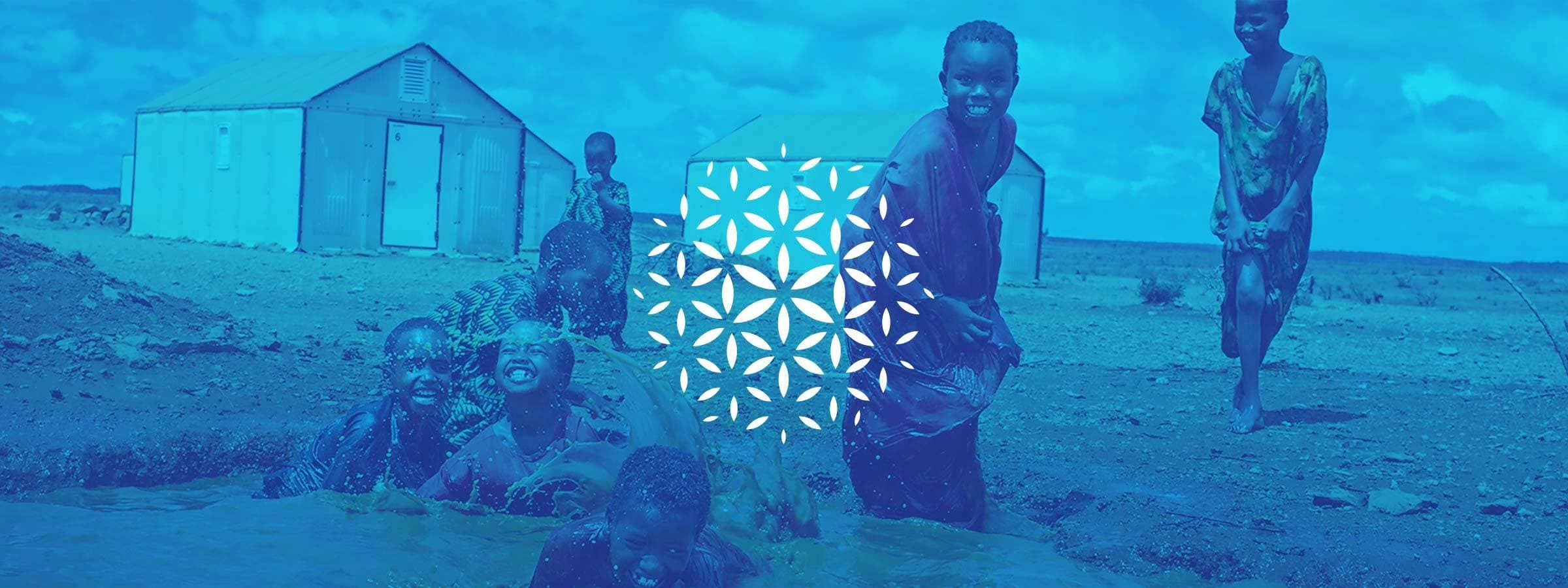 A group of smiling children play in a body of water in a rural area. Simple buildings are in the background, and the image features a superimposed geometric pattern. The photo is tinted blue.