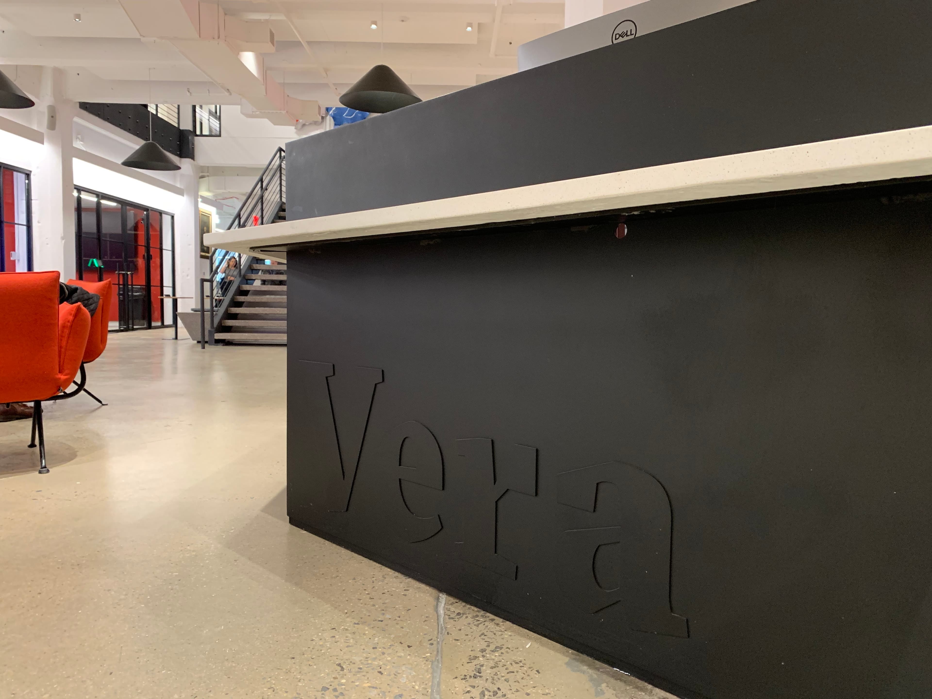 A modern office reception area featuring a sleek black desk with the word "Vera" embossed on it. To the left, there's a red couch and glass walls, with a black staircase in the background. The floor is polished concrete, and pendant lights hang from the ceiling.
