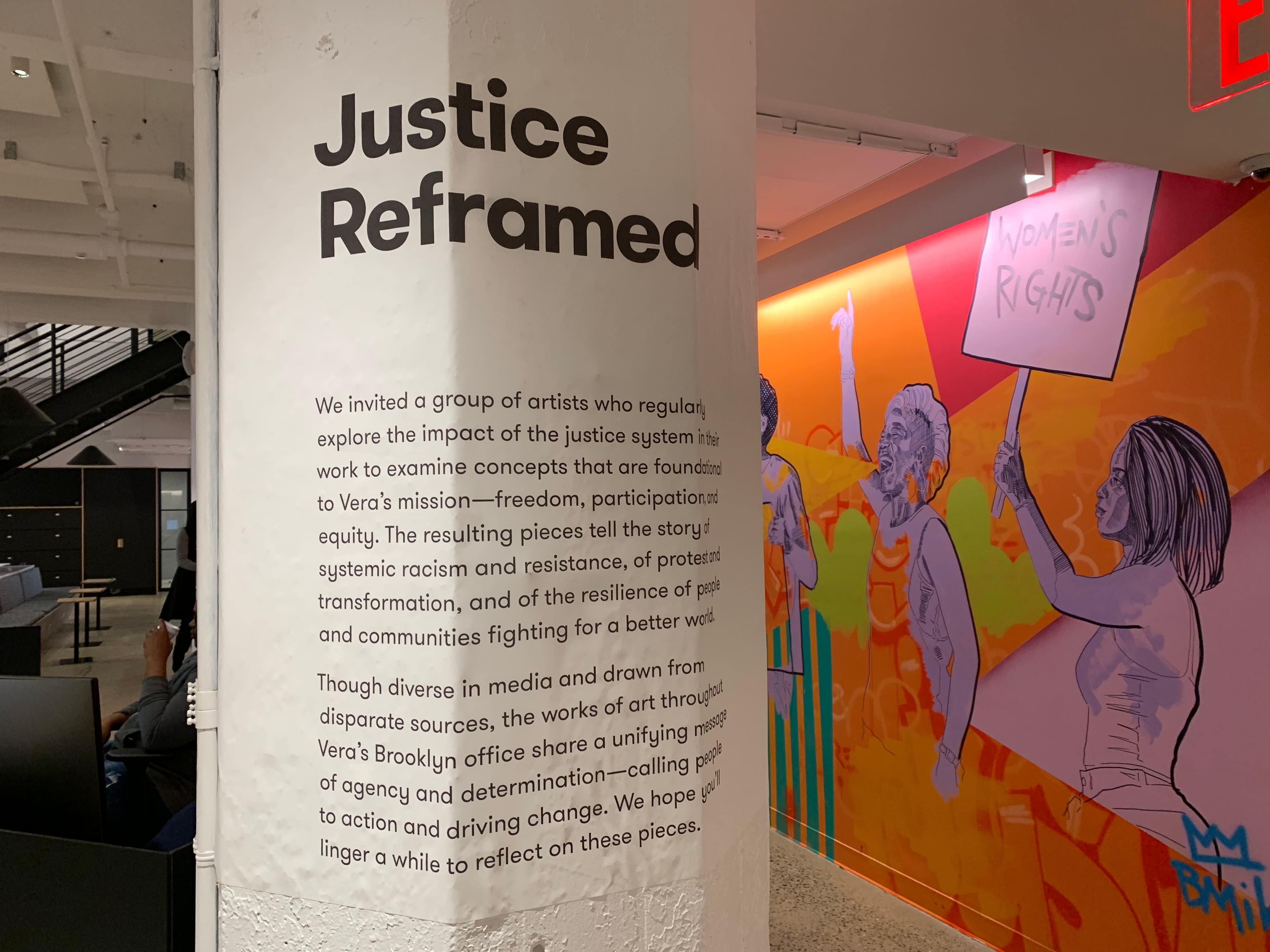 A white pillar displays text discussing the exploration of justice through art, part of Vera's mission. In the background, there is a mural of diverse individuals protesting with signs, including one that reads "Women's Rights," emphasizing social activism.