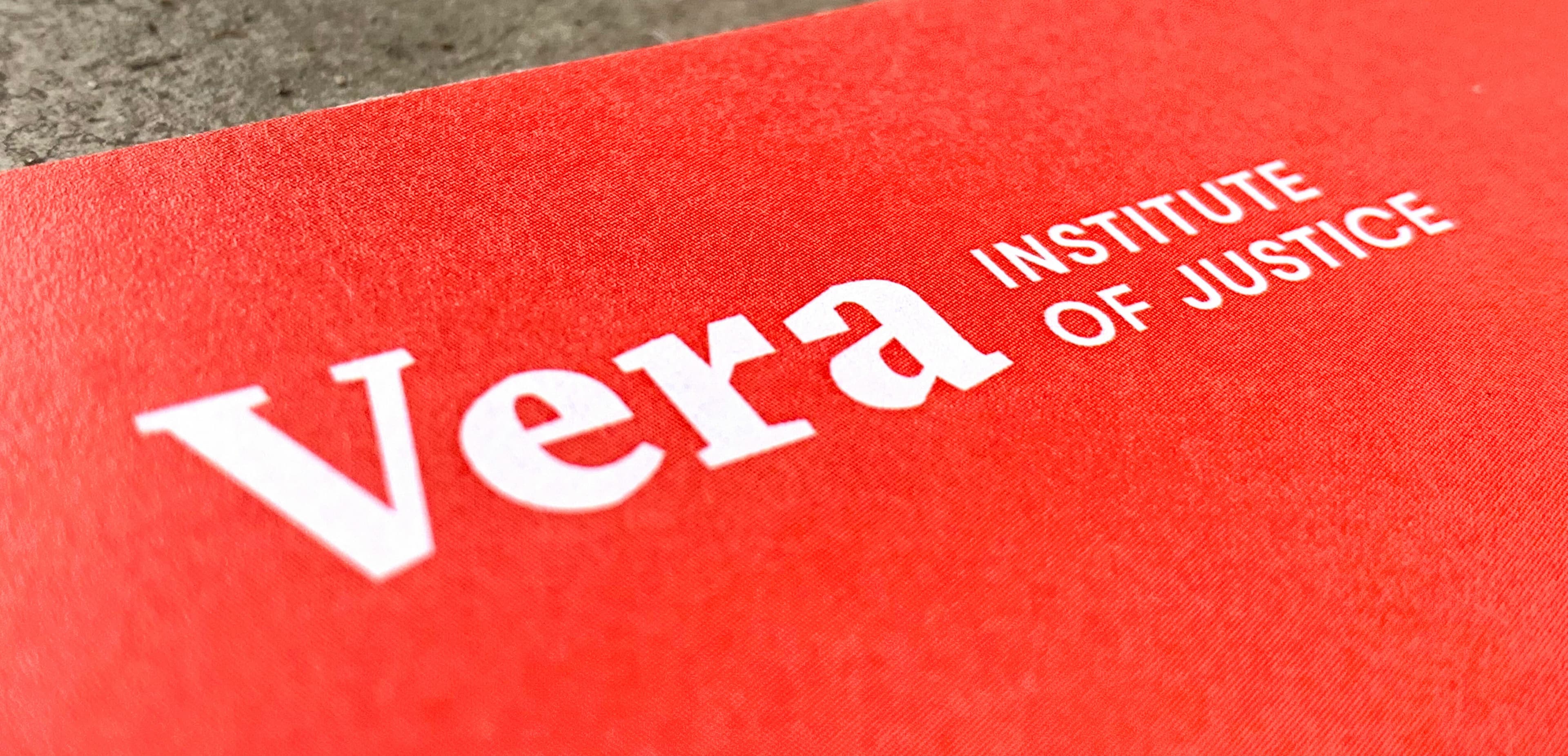 Close-up of a bright red document cover with the white text “Vera Institute of Justice” prominently displayed. The background appears textured, suggesting the document rests on a rough surface.