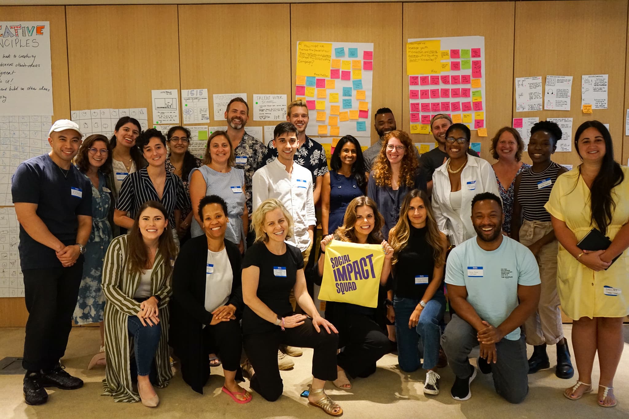 A diverse group of around 25 people smiling and posing for a group photo indoors. Some are seated while others stand behind them. One person holds a yellow flag with the text "Social Impact Squad." Colorful post-it notes and charts cover the wall in the background.