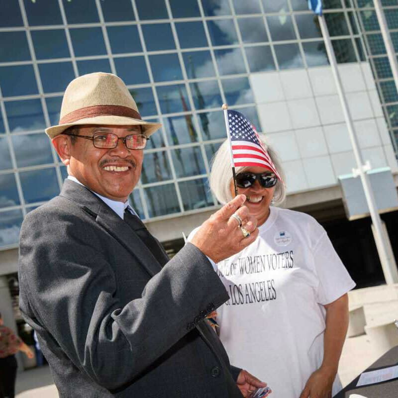 A man wearing a hat and suit holds a small American flag, smiling at the camera. Beside him, a woman with blonde hair wearing a white T-shirt and sunglasses also smiles. They stand in front of a glass building under a bright sky.