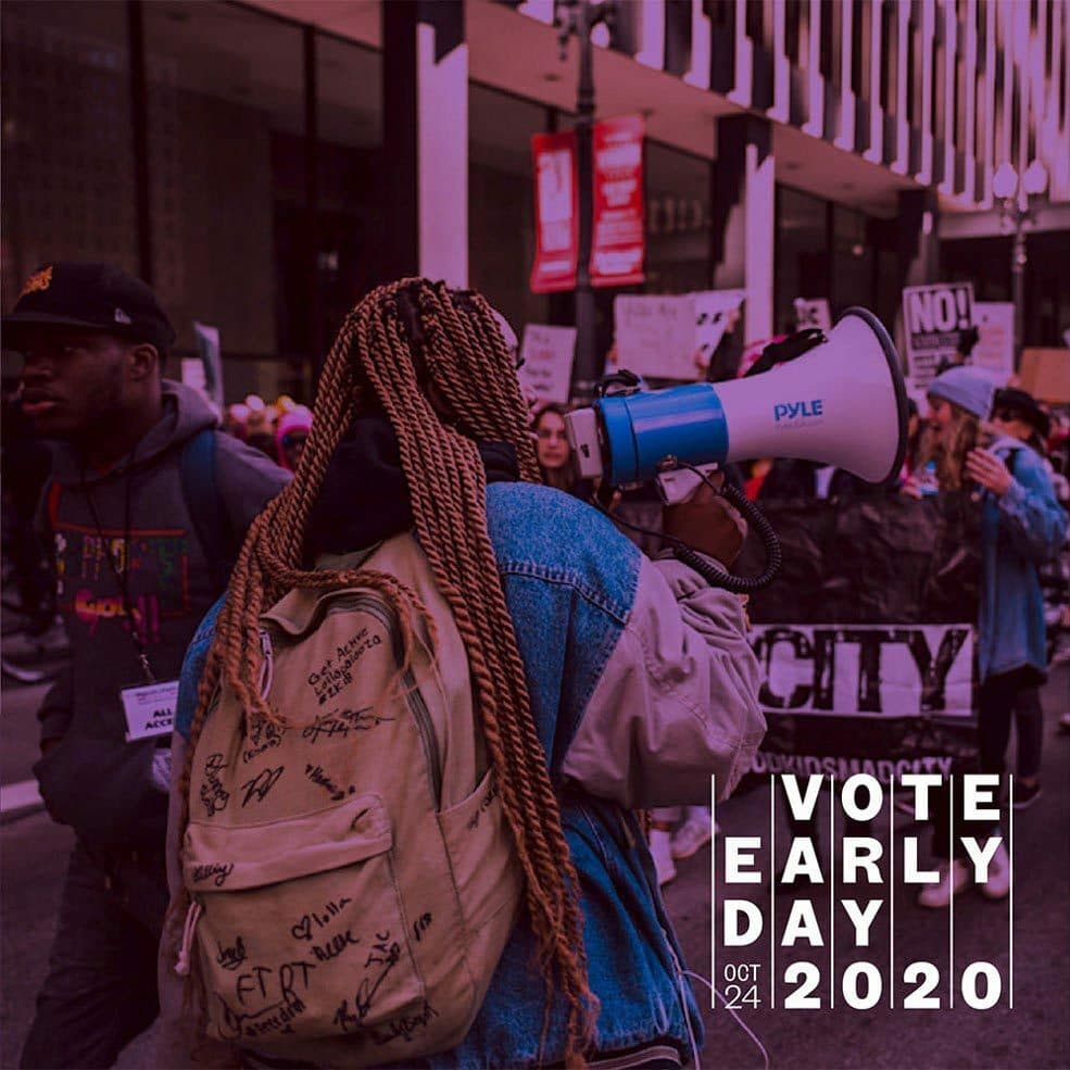 A person with long braids holding a blue and white megaphone is seen from behind, addressing a crowd at a demonstration. They are wearing a denim jacket and a beige backpack. The image features the text "VOTE EARLY DAY OCT 24 2020" in white letters at the bottom right.