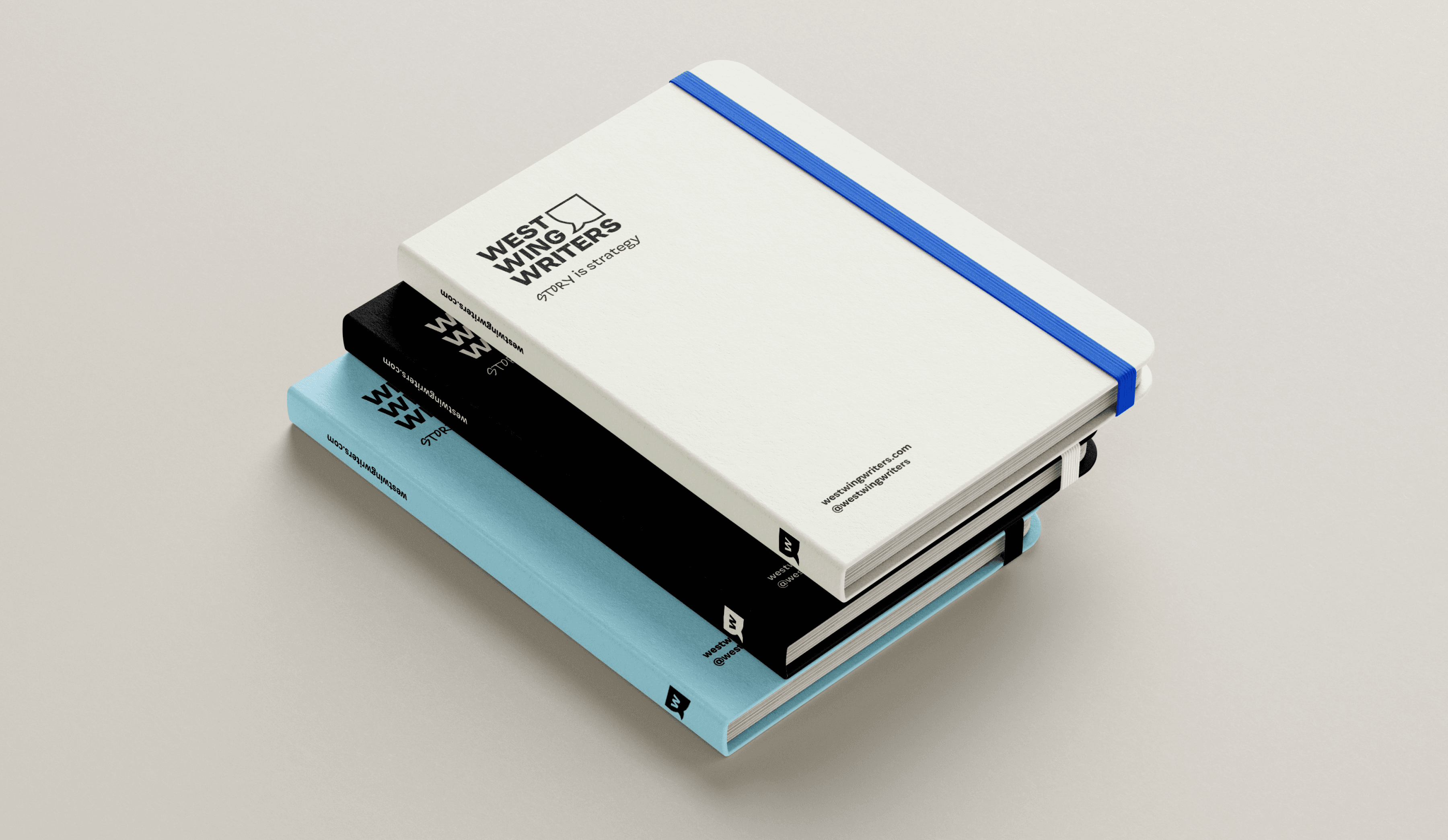 A stack of three notebooks with different colored covers: the top notebook is white with a blue elastic band, the middle one is black, and the bottom one is light blue. Each notebook features the logo and text “West Wing Writers.”.