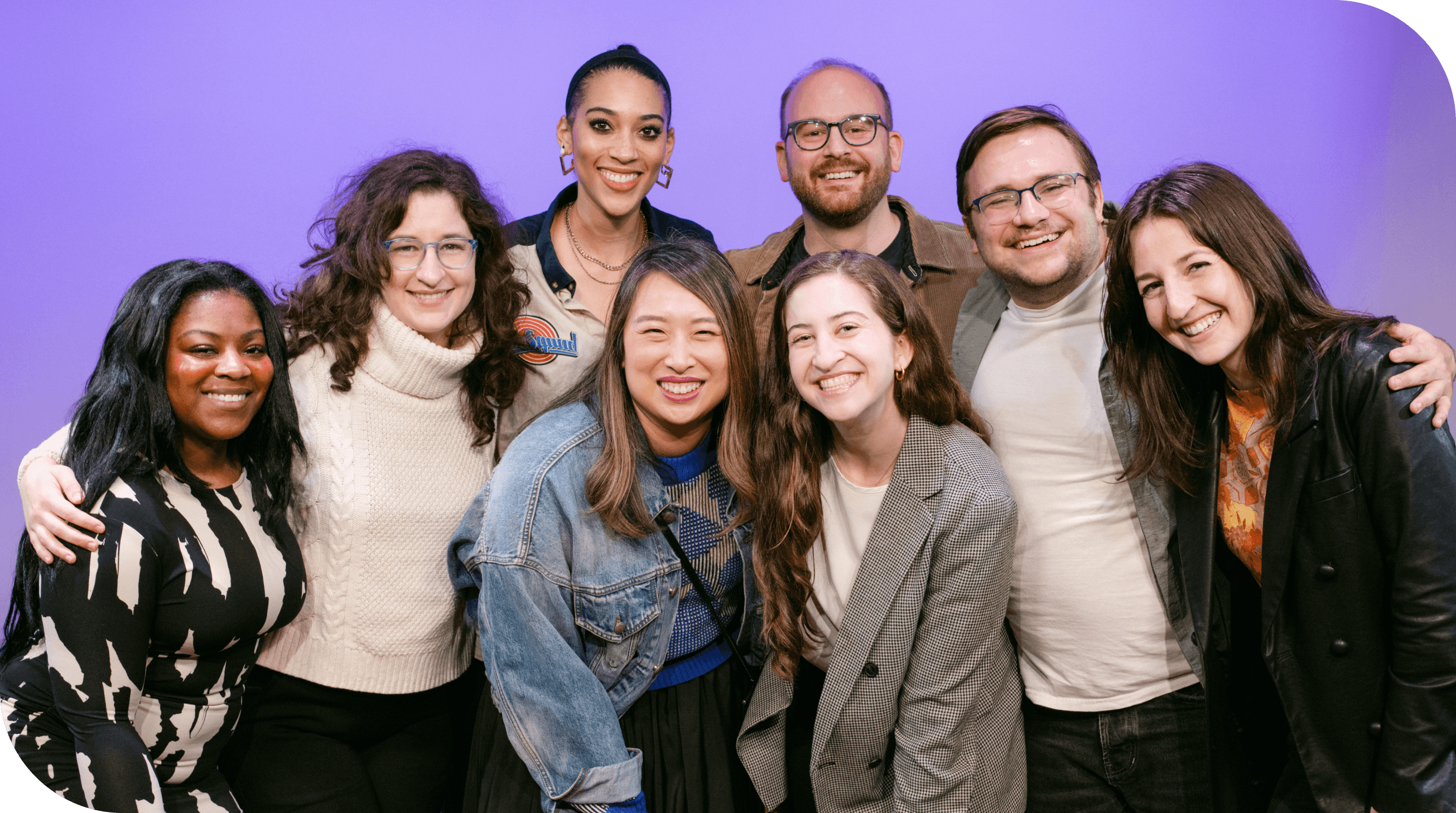 A group of eight people, composed of six women and two men, are standing together and smiling at the camera against a purple background. They appear happy and are wearing casual to semi-casual attire, with some people wearing jackets and sweaters.