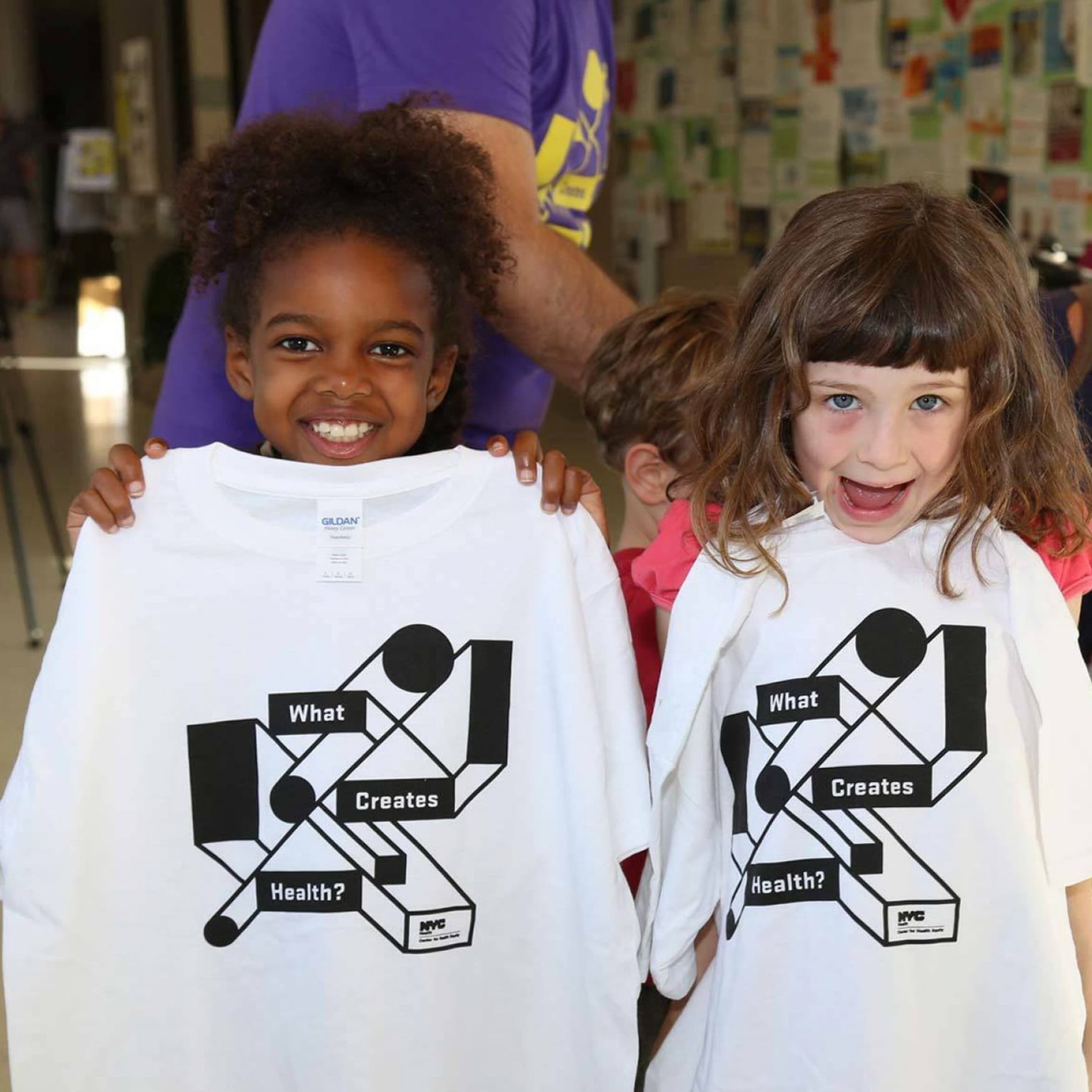 Two young girls smile and hold up white T-shirts bearing the words "What Creates Health?" in bold black text and geometric designs. They are indoors and a person in a purple shirt is visible behind them. Walls display an array of papers in the background.
