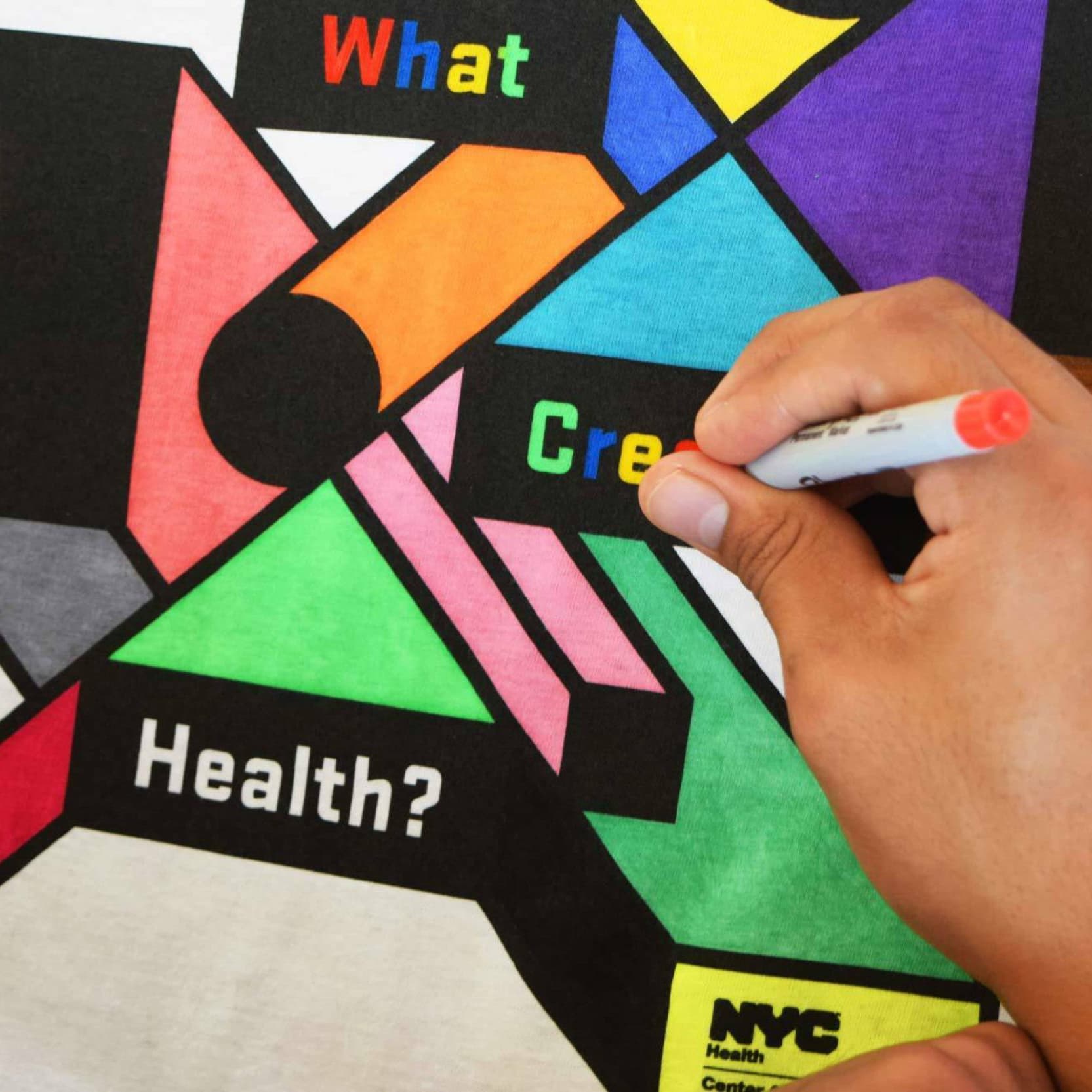 Close-up of a hand using an orange marker to color a brightly colored geometric pattern on a poster. Visible text includes "What," "Health?," "Cre," and "NYC Health." The poster features various interconnected shapes in different colors.