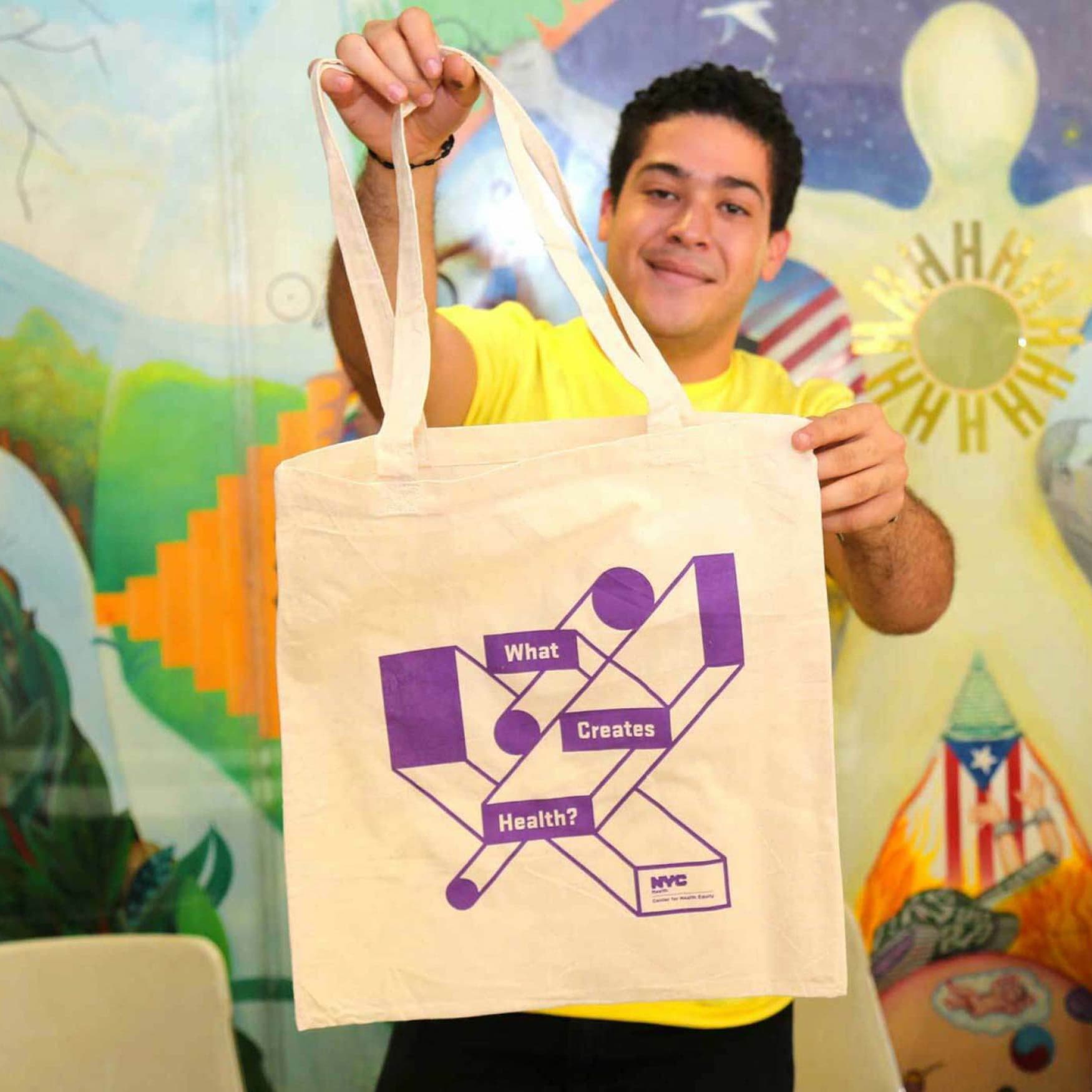 A person wearing a yellow shirt holds up a beige tote bag with purple geometric designs and text reading "What Creates Health?" The background features a colorful mural with abstract shapes and symbols, including a sun and a flag-like element.