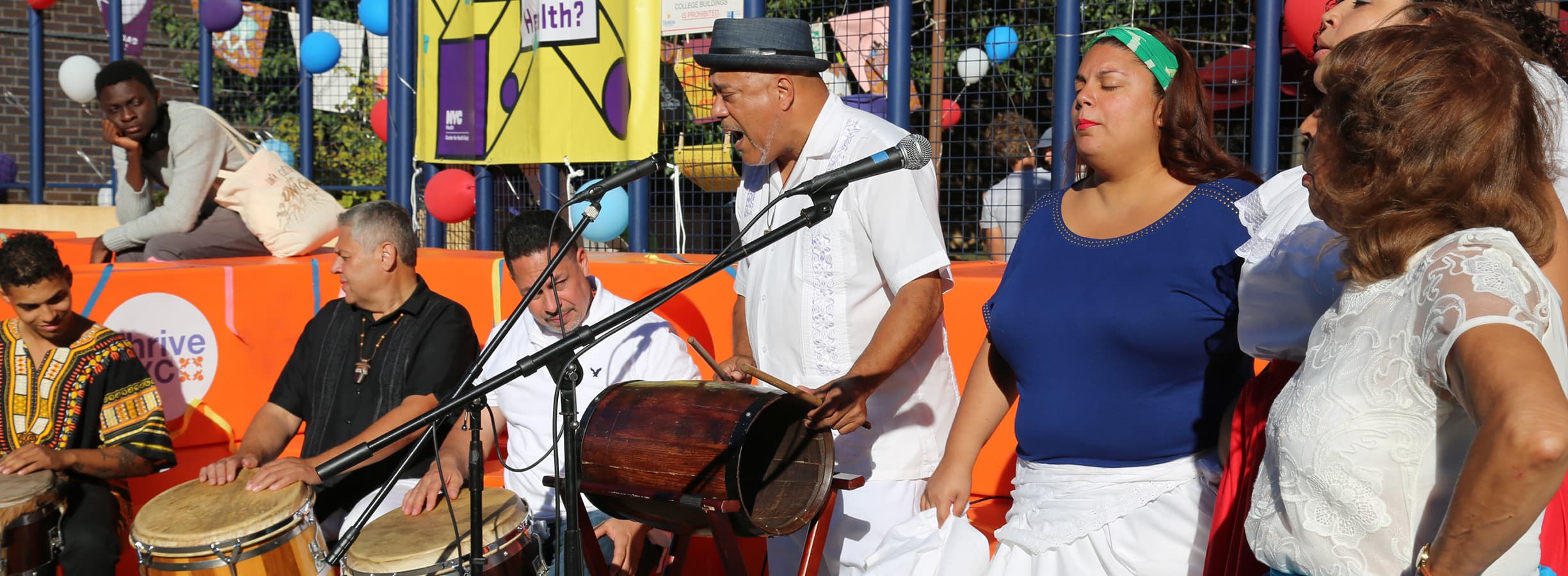 A group of people engaged in a musical performance outdoors. One person is playing a drum, another is seated with bongos, and others are singing. Colorful balloons and banners decorate the background, and an audience listens attentively. Bright, sunny day.