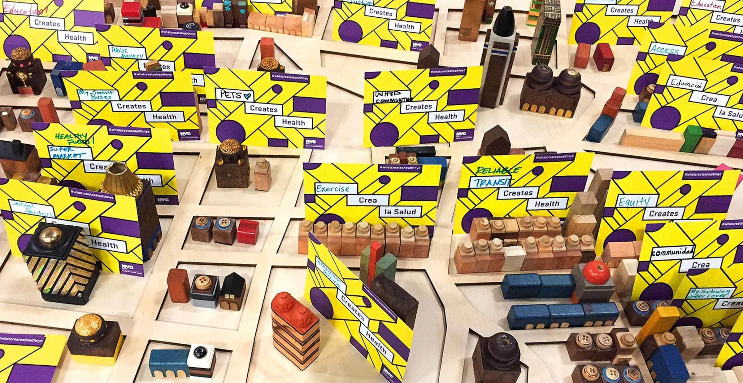 A table filled with numerous colorful building blocks and mini-structures. Bright yellow and purple cards with text such as "Health," "Creates," and "Exercise" are placed amongst the blocks. The scene appears to resemble a playful, educational display.